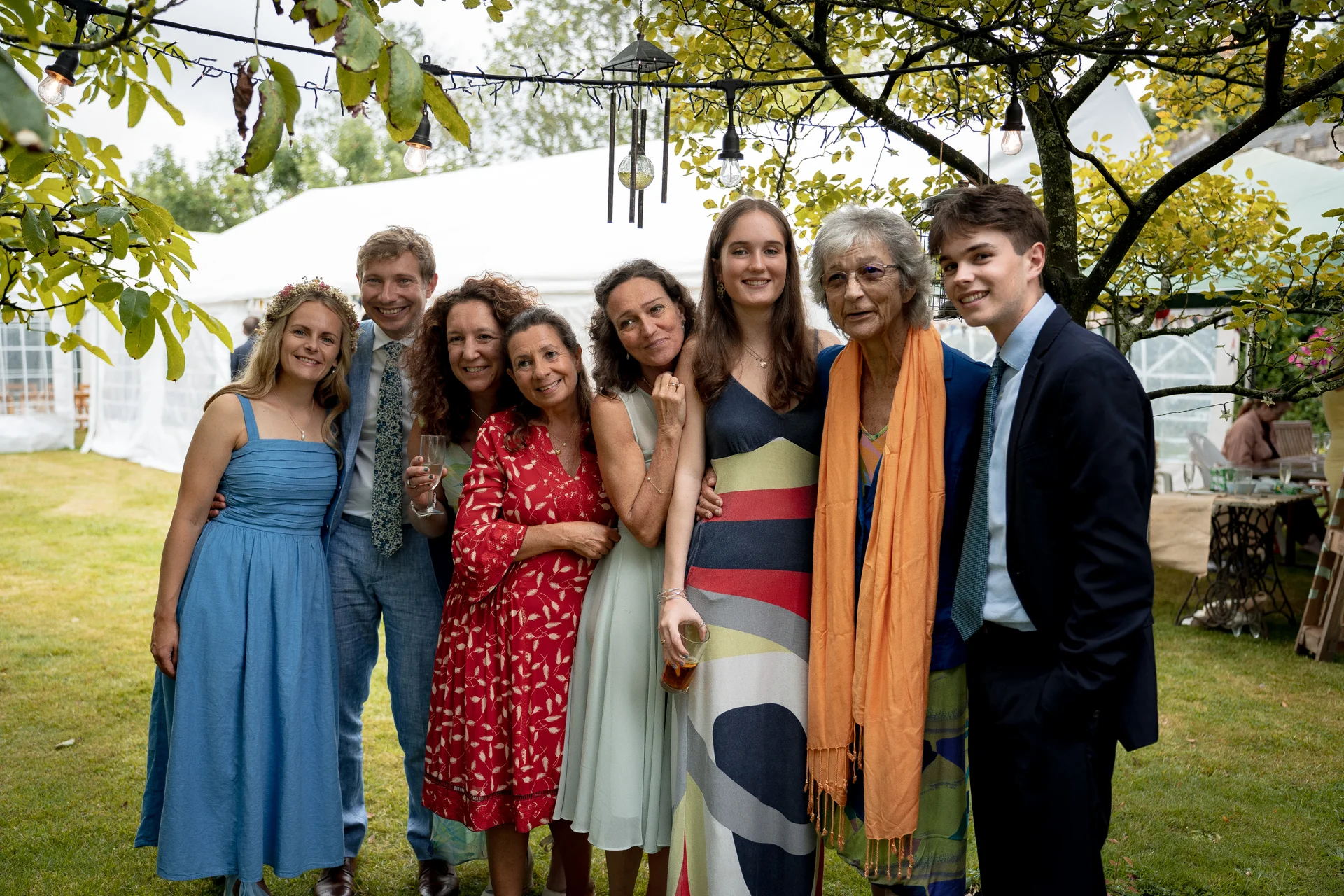A group of family members and wedding guests smile together outdoors in a garden setting beneath festoon lighting and leafy trees.