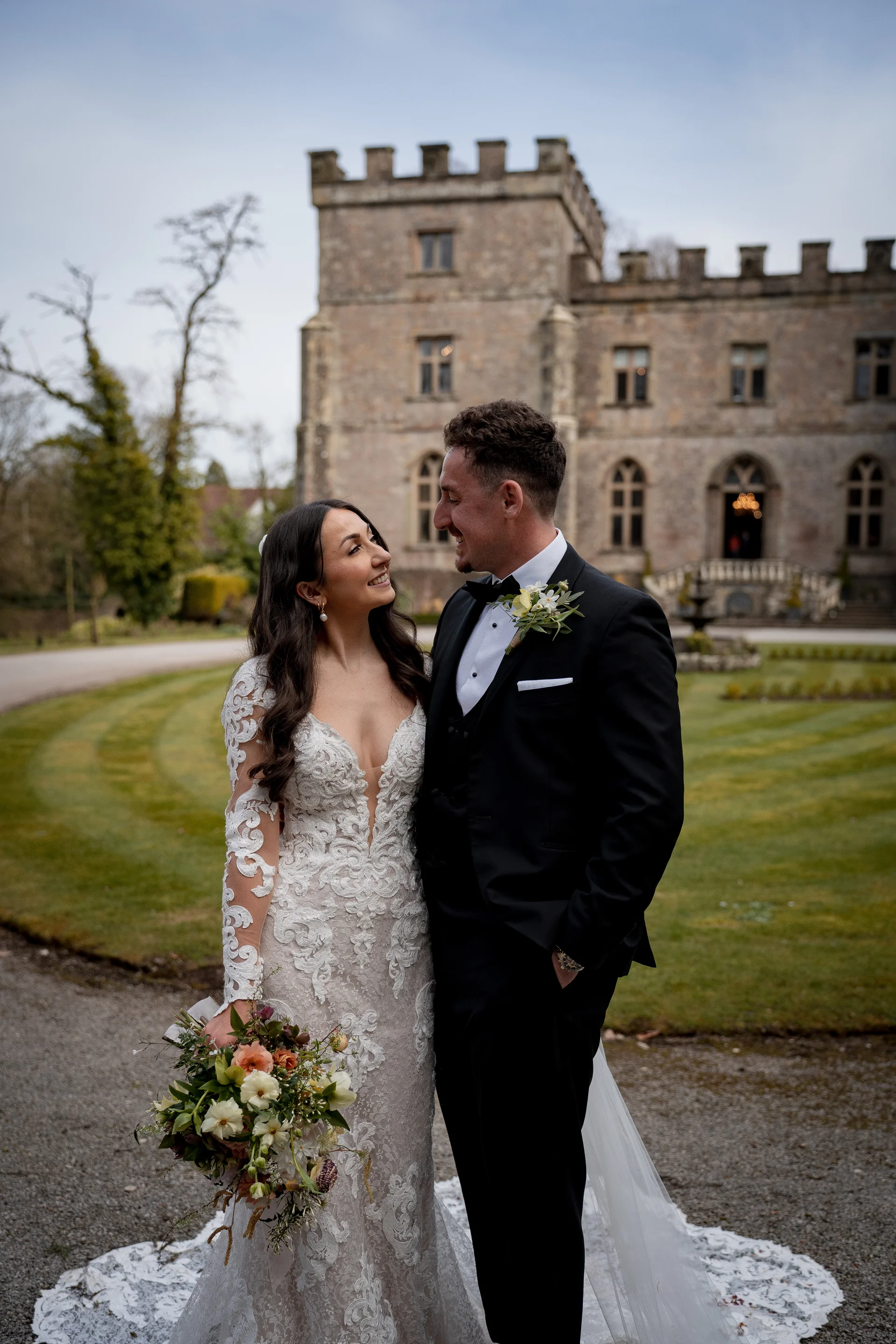 A newlywed couple gaze lovingly at each other outdoors on a gravel pathway with a grand stone castle and manicured lawn in the background.