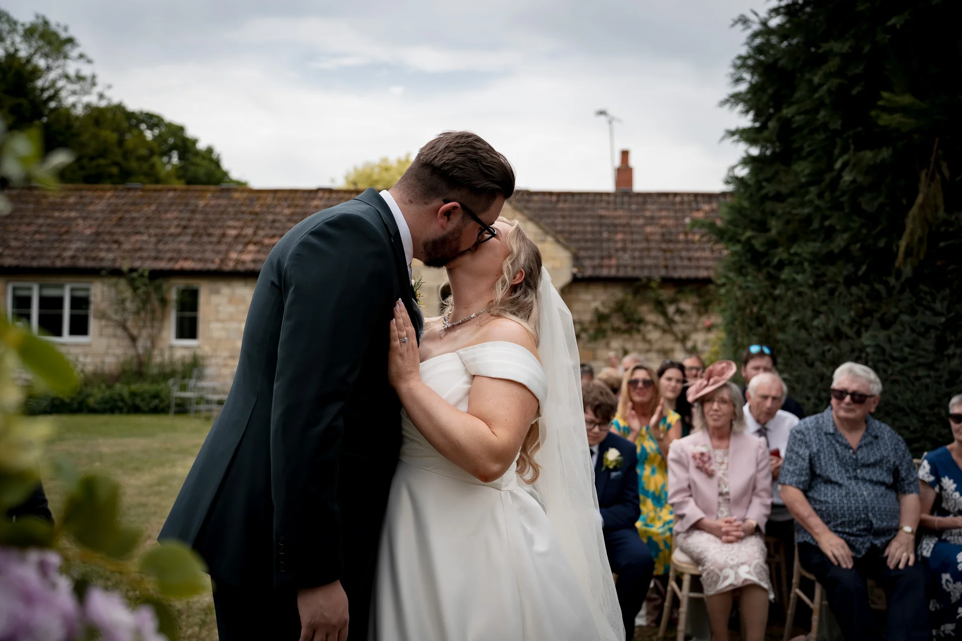 The newly married couple share their first kiss during an outdoor ceremony while guests look on seated in a garden setting.
