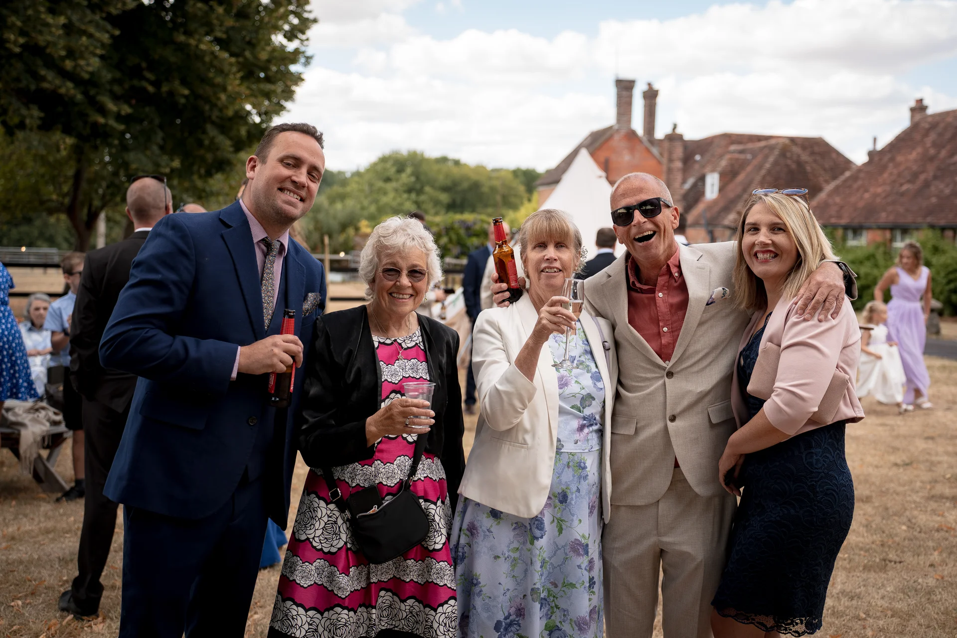 A cheerful group of wedding guests enjoying drinks and laughing together outdoors during a sunny summer reception.