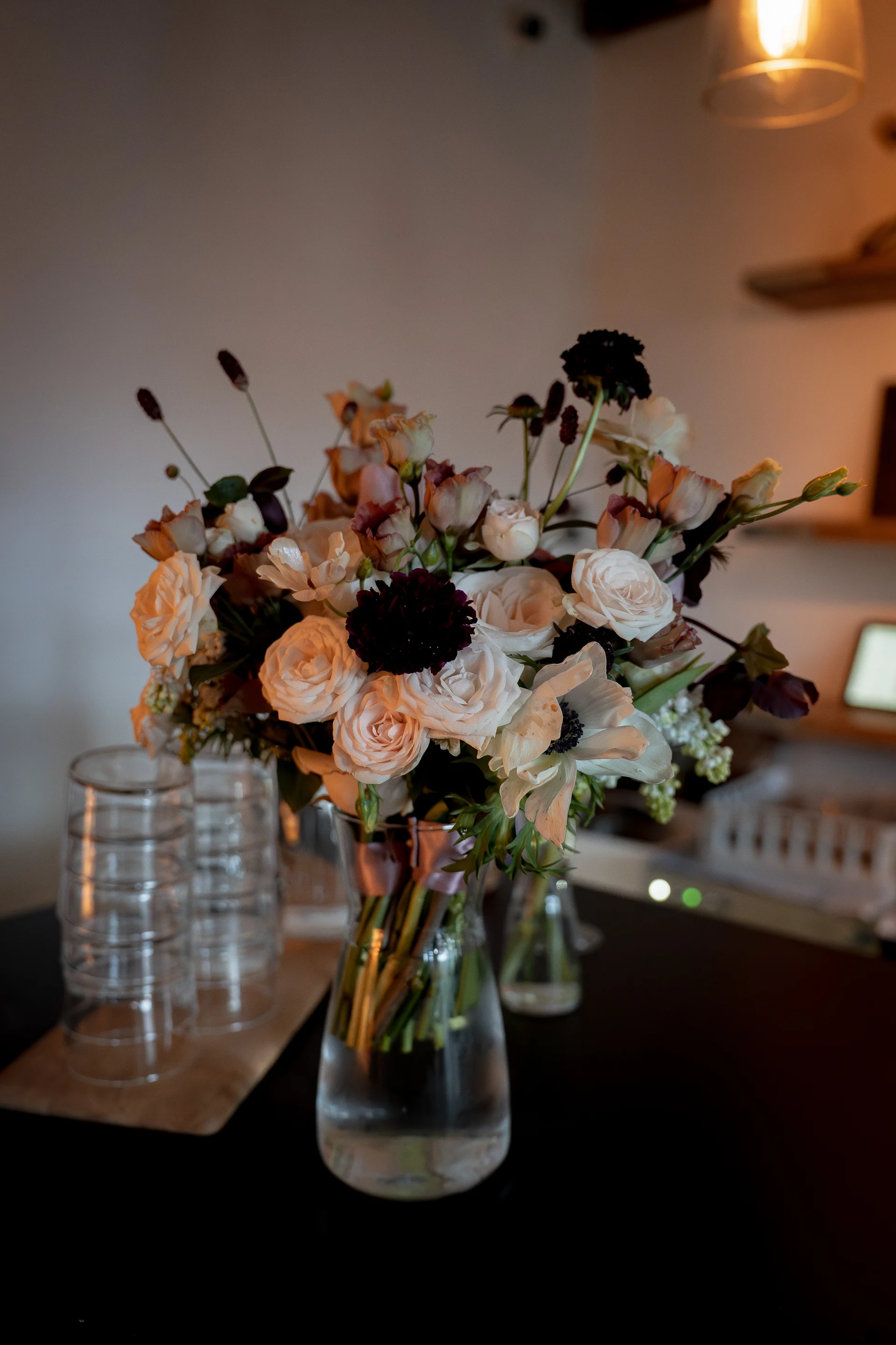 An artfully arranged wedding floral centrepiece featuring blush roses, white anemones, and dark burgundy blooms sits in a clear glass vase on a dark table.