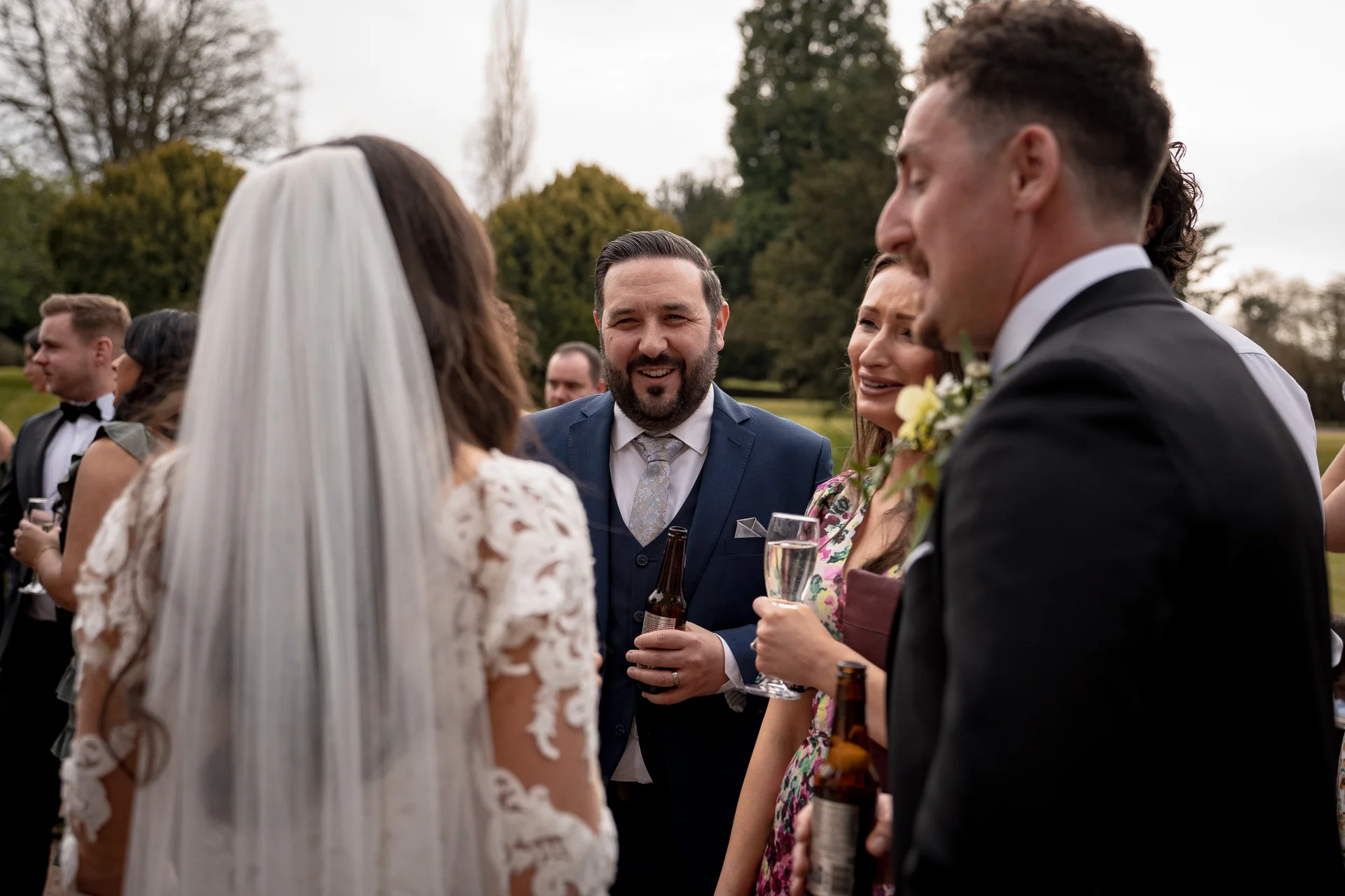 Guests mingle and chat during an outdoor drinks reception while a bride wearing a lace dress and veil stands in the foreground.