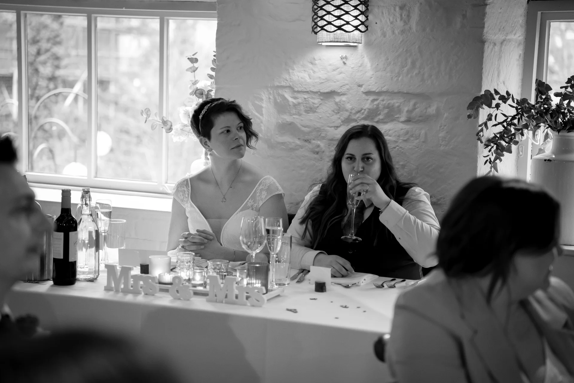 A candid black and white photograph of two brides seated at the top table during wedding speeches, with one bride drinking from a glass.