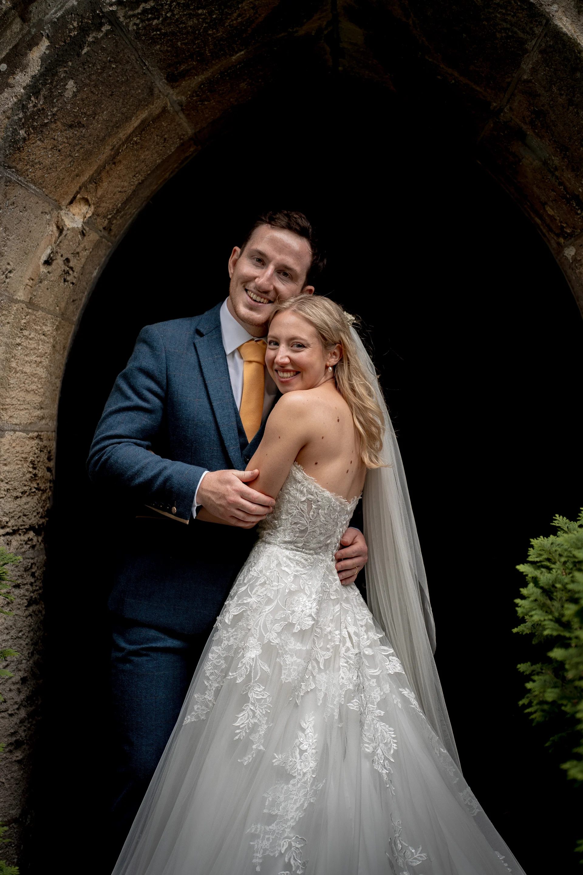 A newlywed couple smiles warmly while embracing inside a historic stone archway after their wedding ceremony.