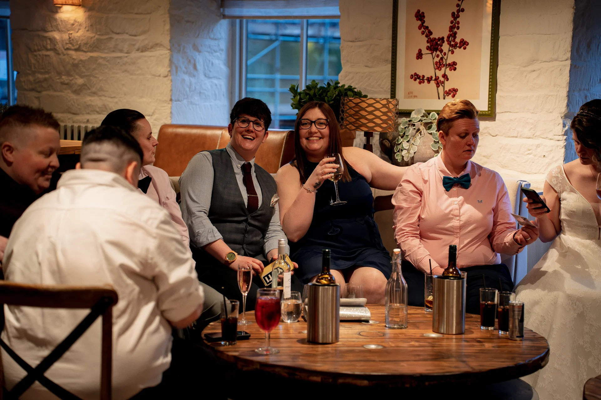 A candid group of wedding guests laughing and relaxing together around a wooden table during the drinks reception in a pub venue.