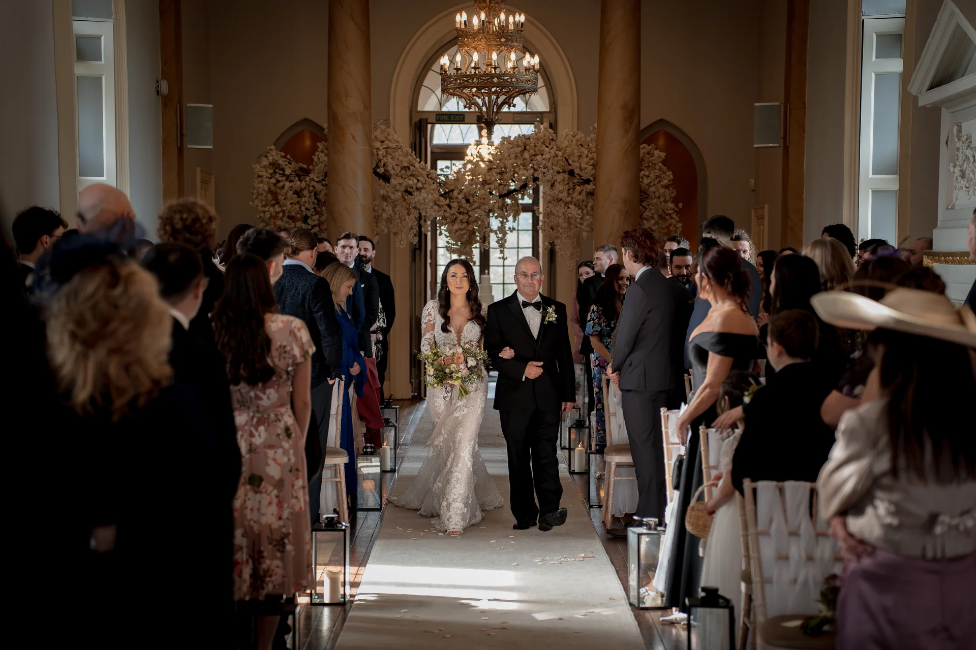 A bride walks down the aisle arm in arm with her father in a grand stately home ceremony space decorated with a large floral arch.