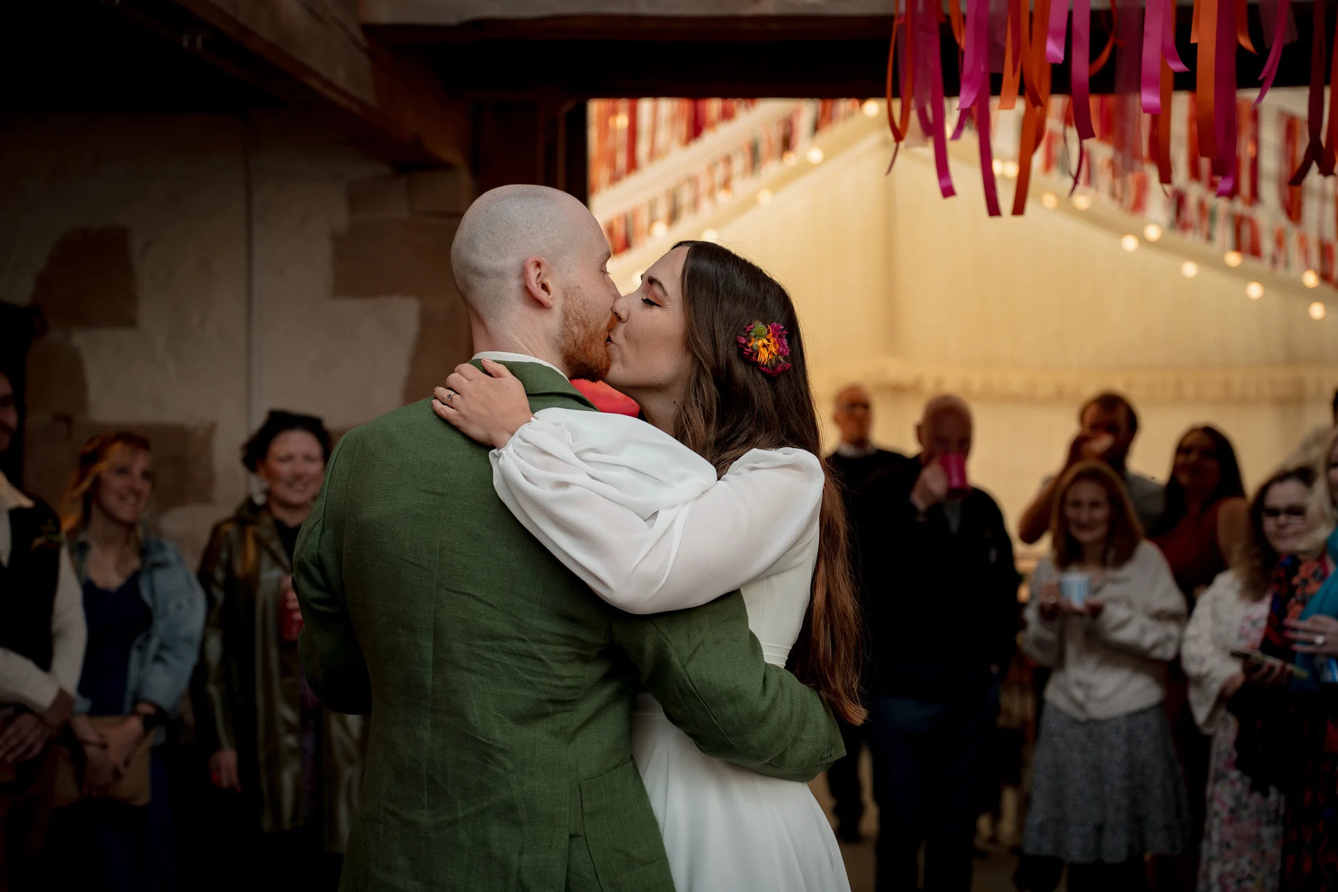 Newlyweds share an intimate kiss during their first dance inside a rustic barn decorated with colourful hanging ribbons while guests watch on happily.