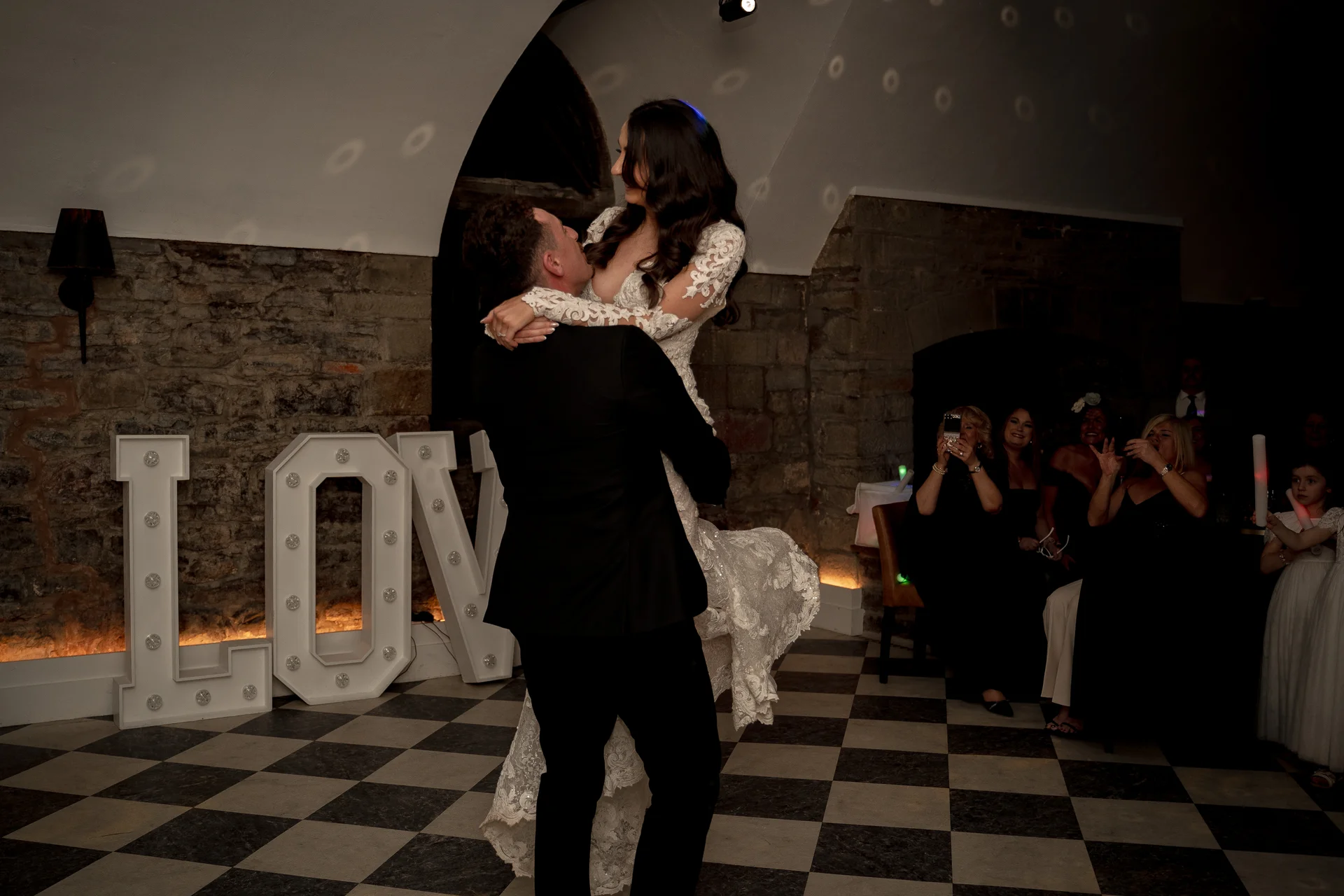 The groom lifts the bride into the air during their first dance on a checkered dance floor in front of illuminated light up letters.