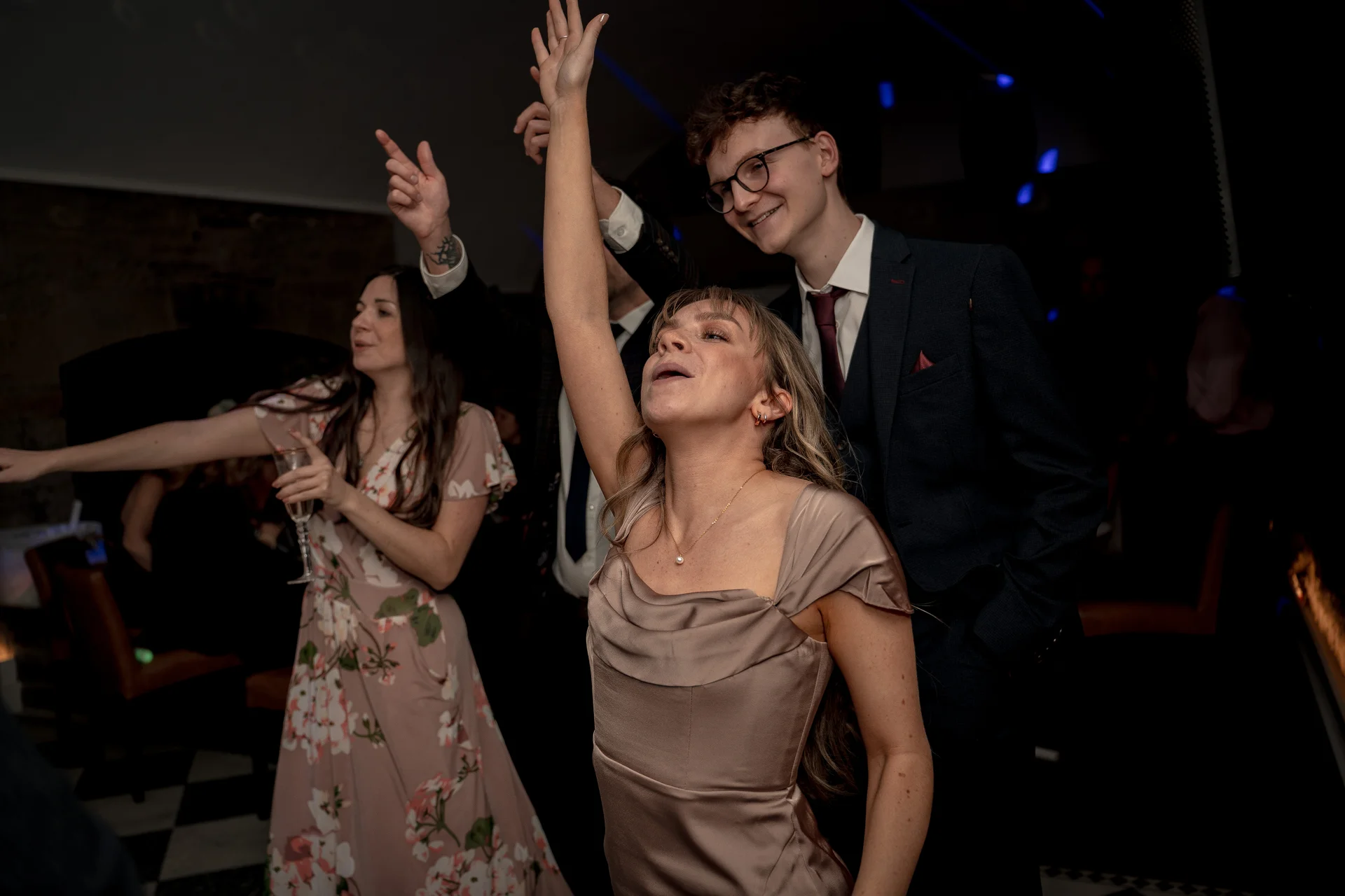 Wedding guests enjoy dancing enthusiastically on the dark reception floor with arms raised and joyful expressions.