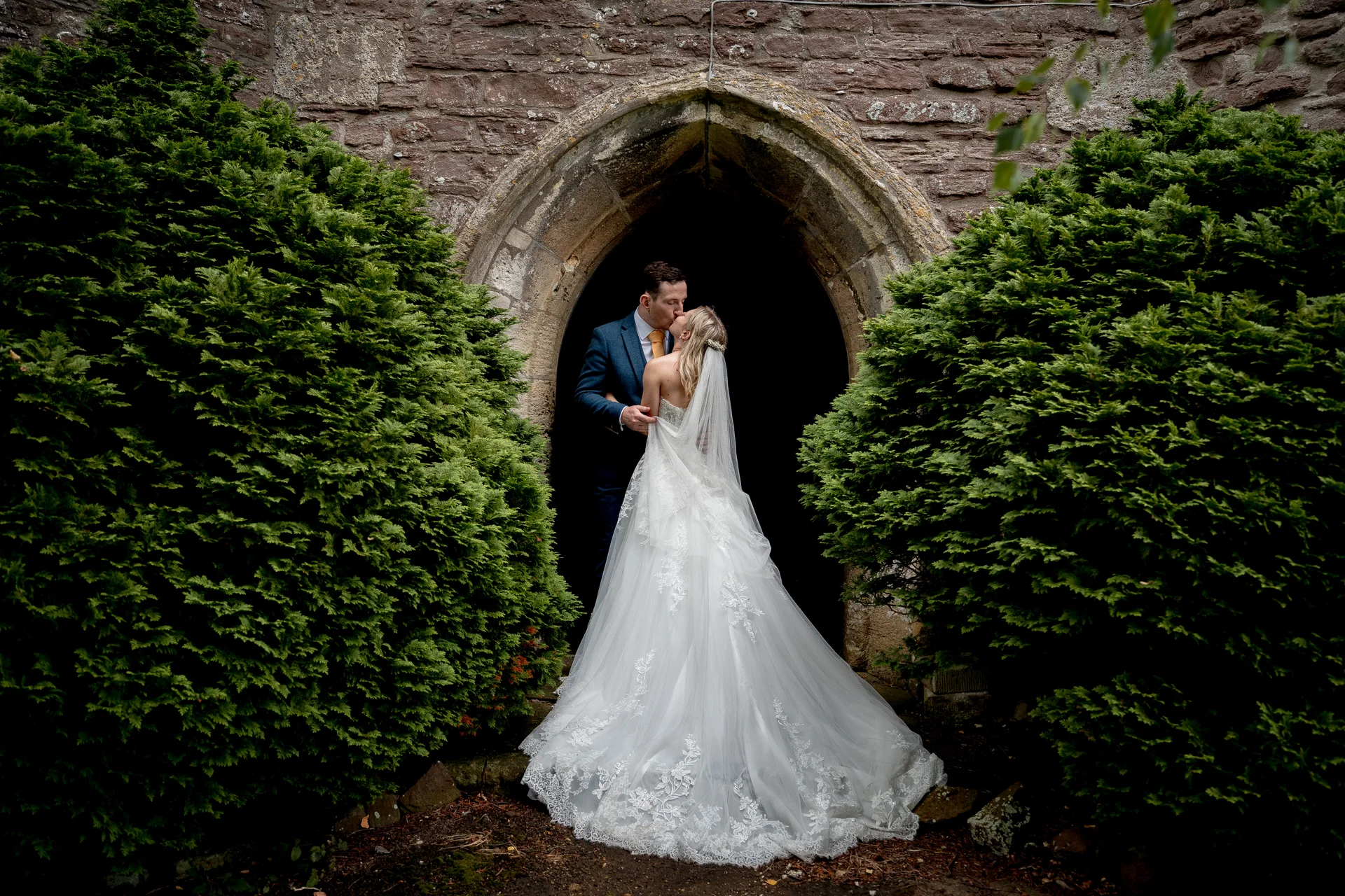 A newlywed couple shares a tender kiss framed by an ancient stone church archway flanked by large green topiary bushes.