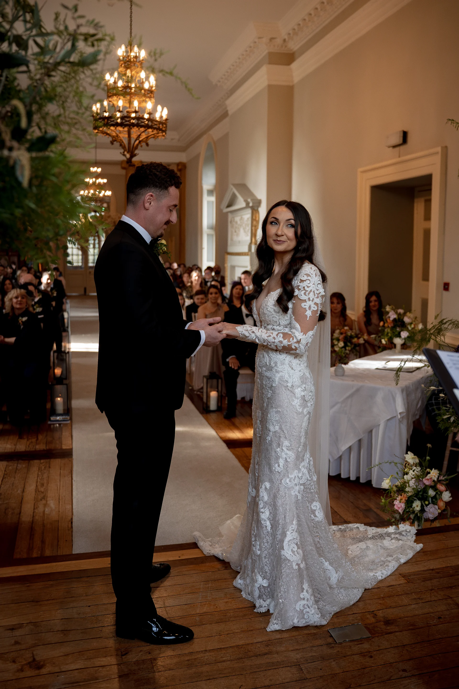 The bride and groom exchange rings during their indoor wedding ceremony inside an elegant ballroom lit by ornate chandeliers.