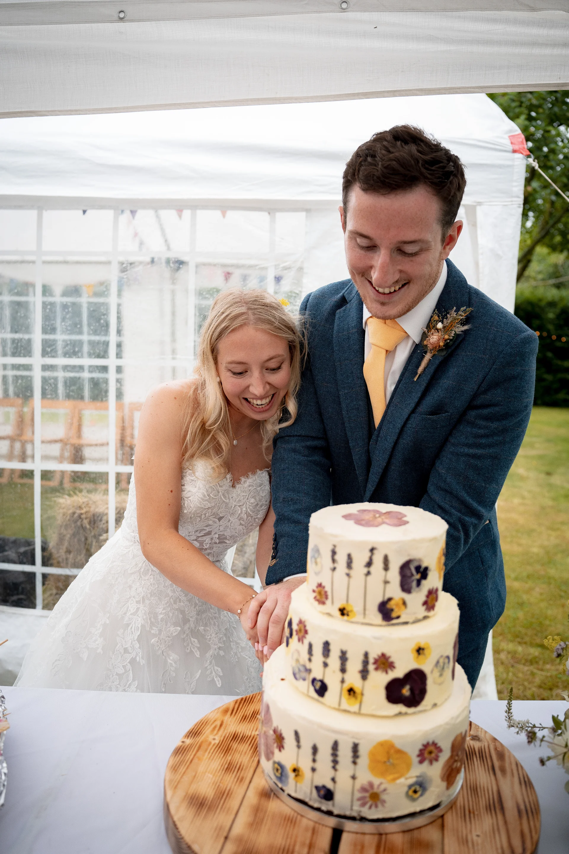 A smiling bride and groom cut into a three-tiered wedding cake decorated with pressed flowers inside a white marquee.