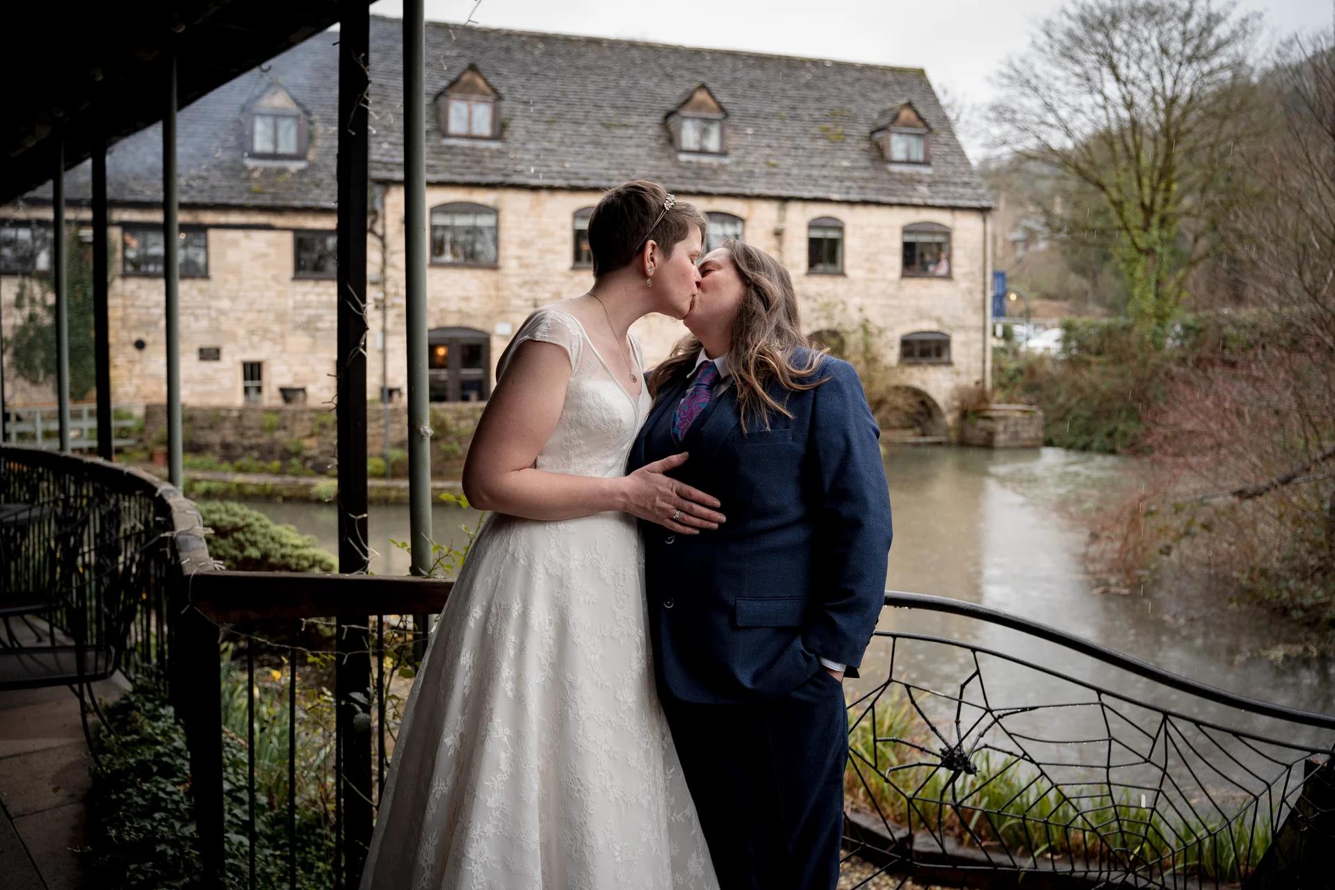 Two brides share a tender kiss on a covered terrace overlooking a historic stone mill and river on an overcast wedding day.