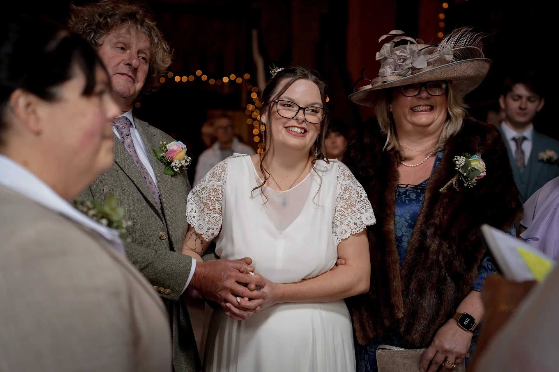 A smiling bride holding hands during her wedding ceremony, surrounded by family members and guests in a warm indoor setting.