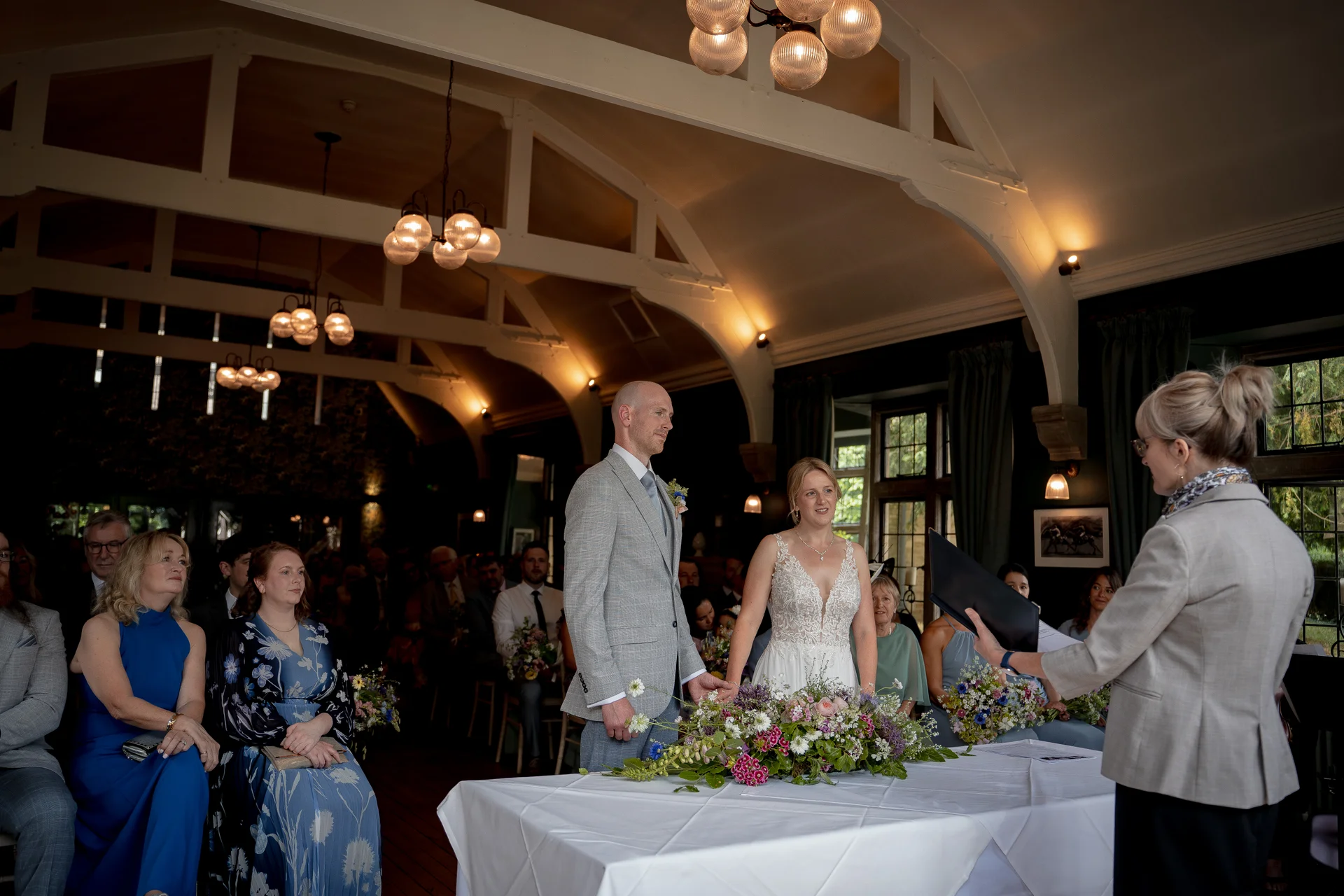A bride in a white gown and a groom in a grey suit stand at a ceremony table before a celebrant during their wedding.