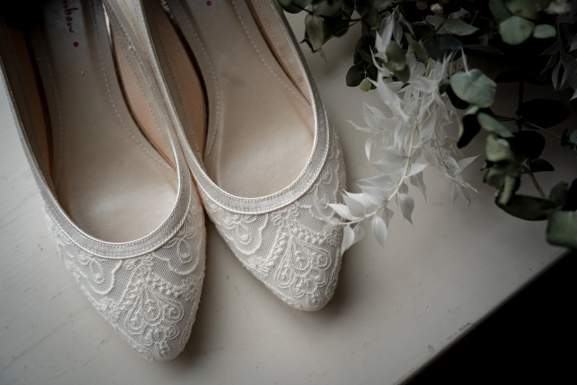 An elegant flat lay detail of ivory lace bridal shoes paired with delicate green and white foliage.