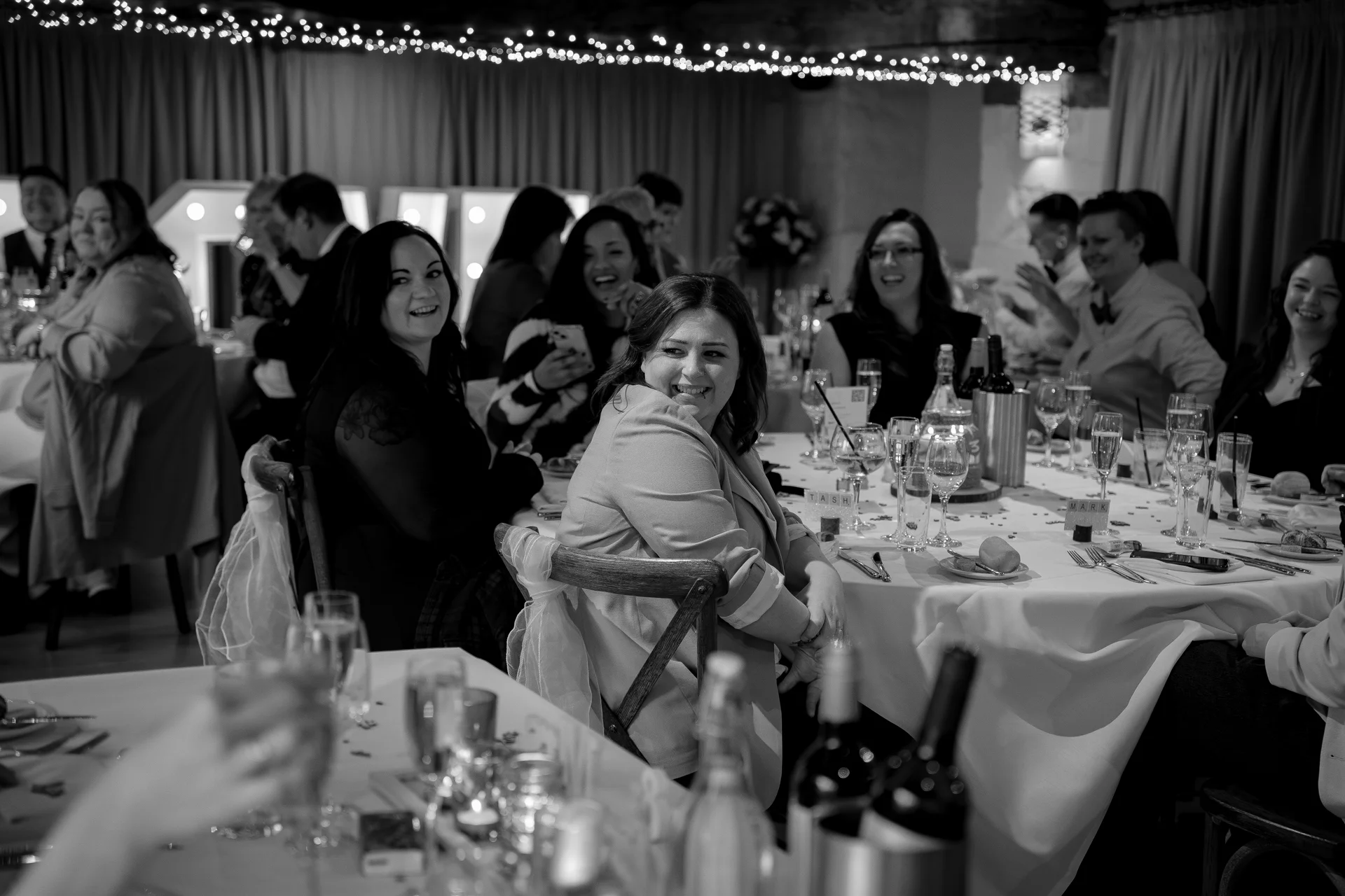 Guests laugh and converse around a banquet table during the wedding reception while seated under a canopy of twinkling fairy lights.