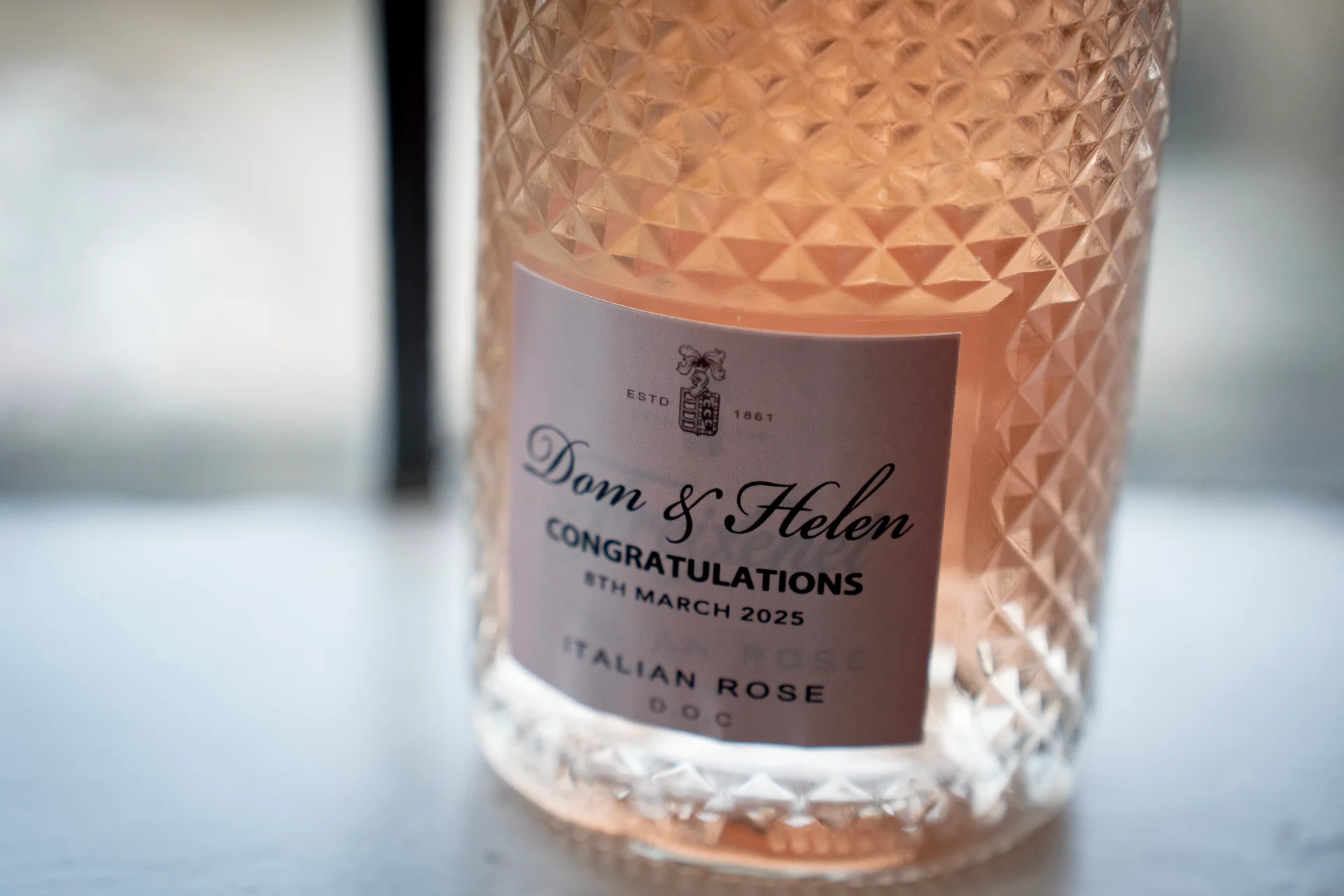 A close-up shot of an ornate crystal glass bottle filled with pink wine, featuring a custom wedding label for Dom and Helen.