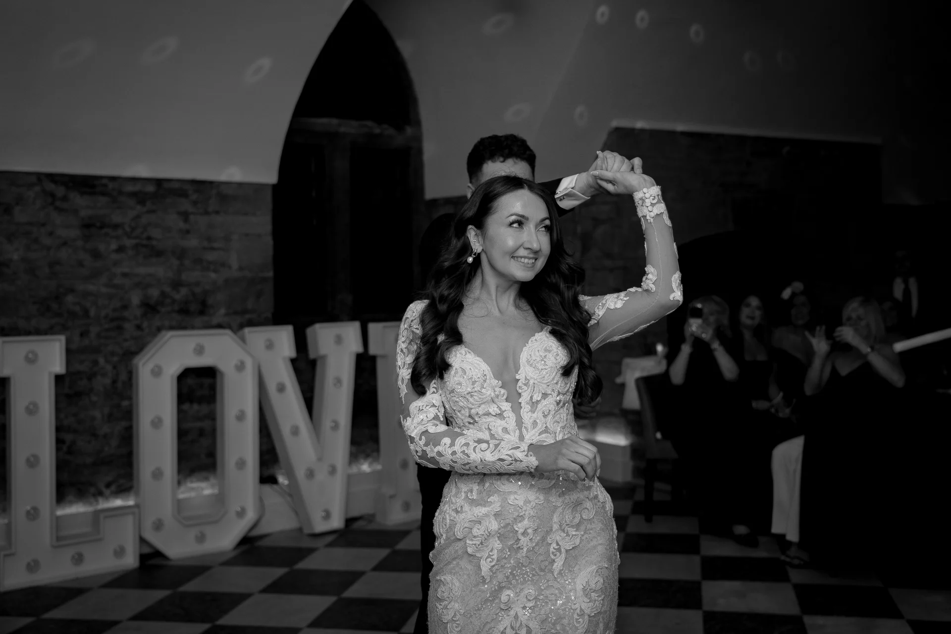 The newlyweds share a joyful first dance together under soft lighting with illuminated marquee letters in the background.