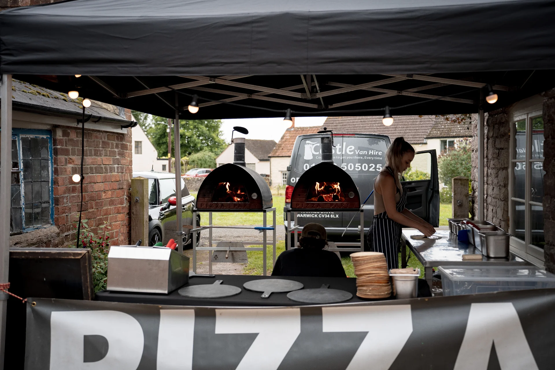 A wedding caterer prepares fresh pizza at an outdoor mobile wood-fired pizza station under a marquee tent.