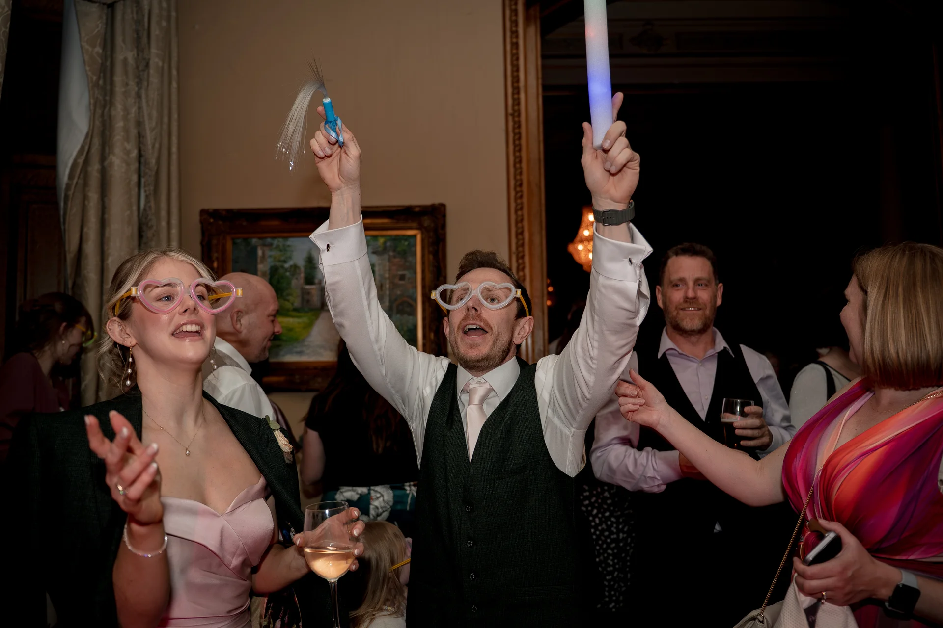 Wedding guests wearing novelty heart-shaped glasses celebrate enthusiastically on the dance floor with arms raised and glowing props.