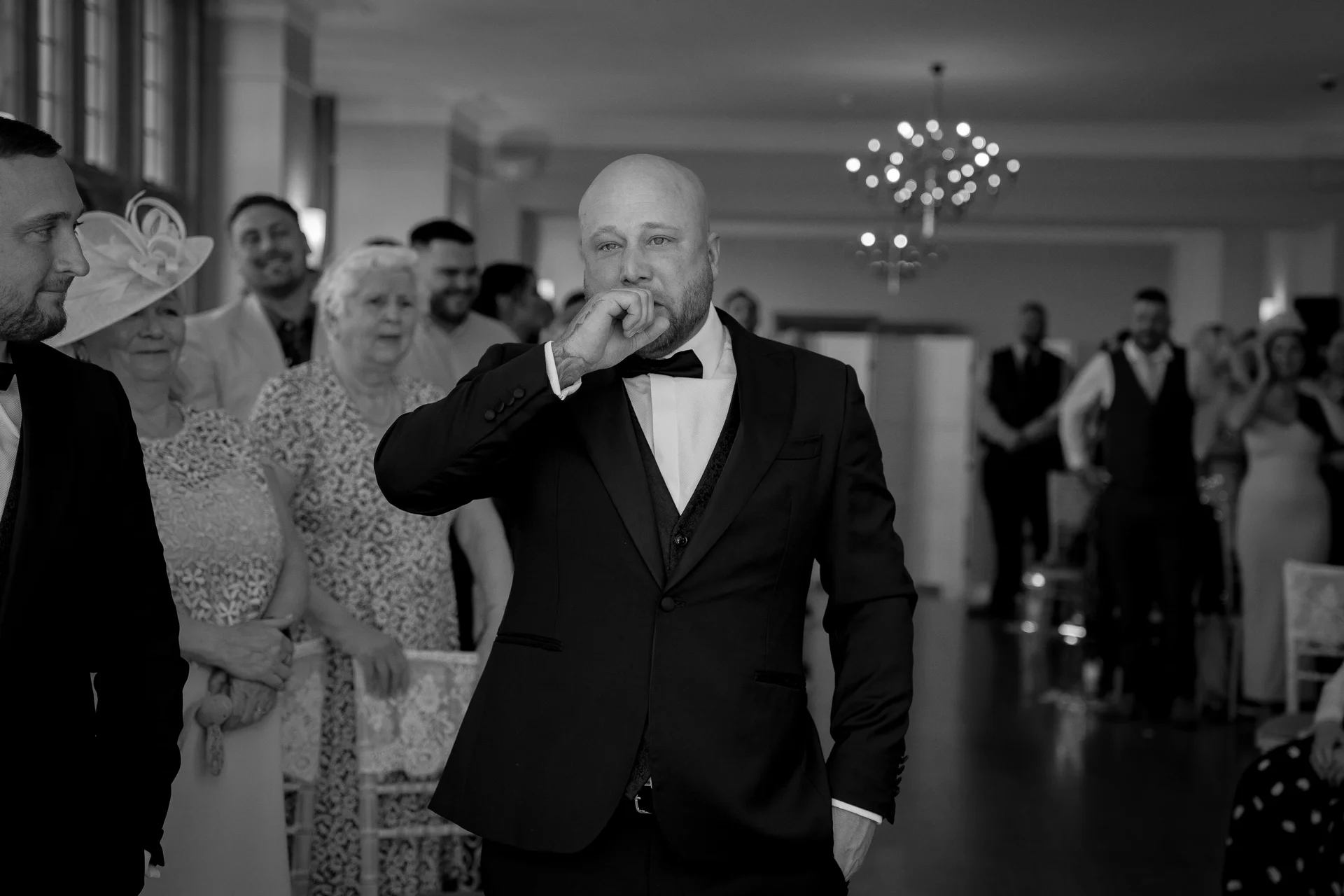An emotional groom in a black tuxedo stands at the altar holding his hand to his mouth with tears in his eyes as wedding guests watch.