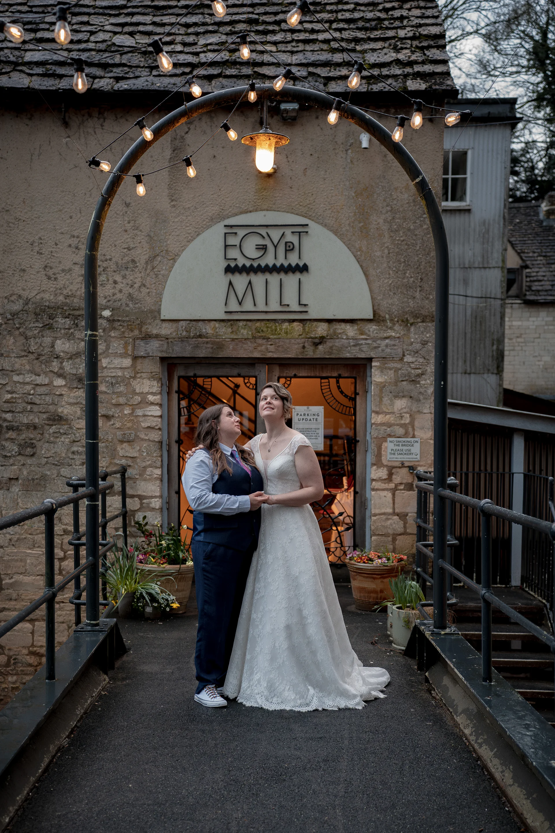 A same-sex couple stands hand in hand on a stone bridge in front of Egypt Mill, looking up toward a glowing arch of string lights above them.