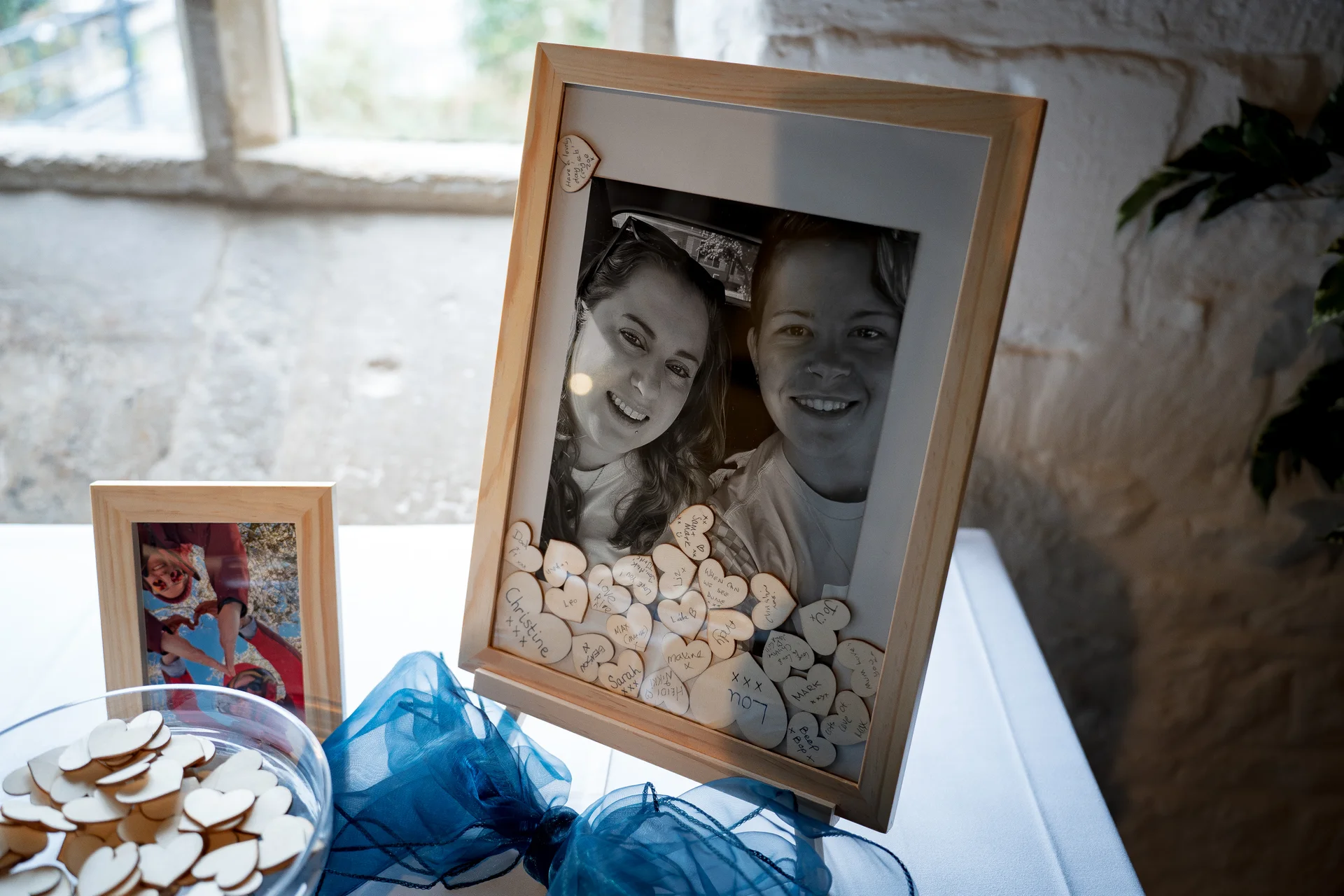 A framed black and white photograph of two smiling brides sits on a table surrounded by signed wooden heart tokens and blue ribbon decor.