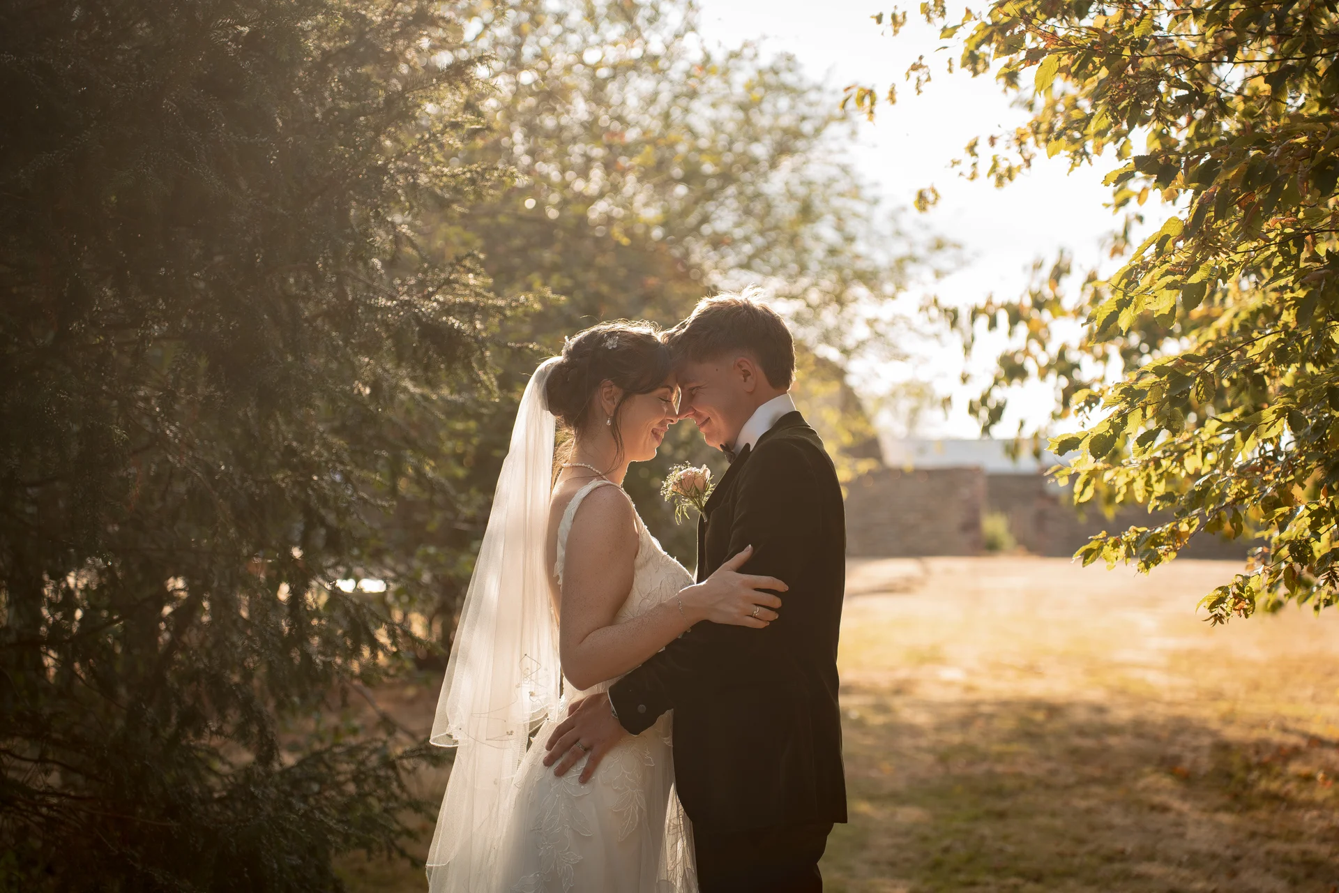 A newlywed couple shares a tender forehead-to-forehead moment outdoors in a sunlit woodland during golden hour, framed by trees and glowing light.