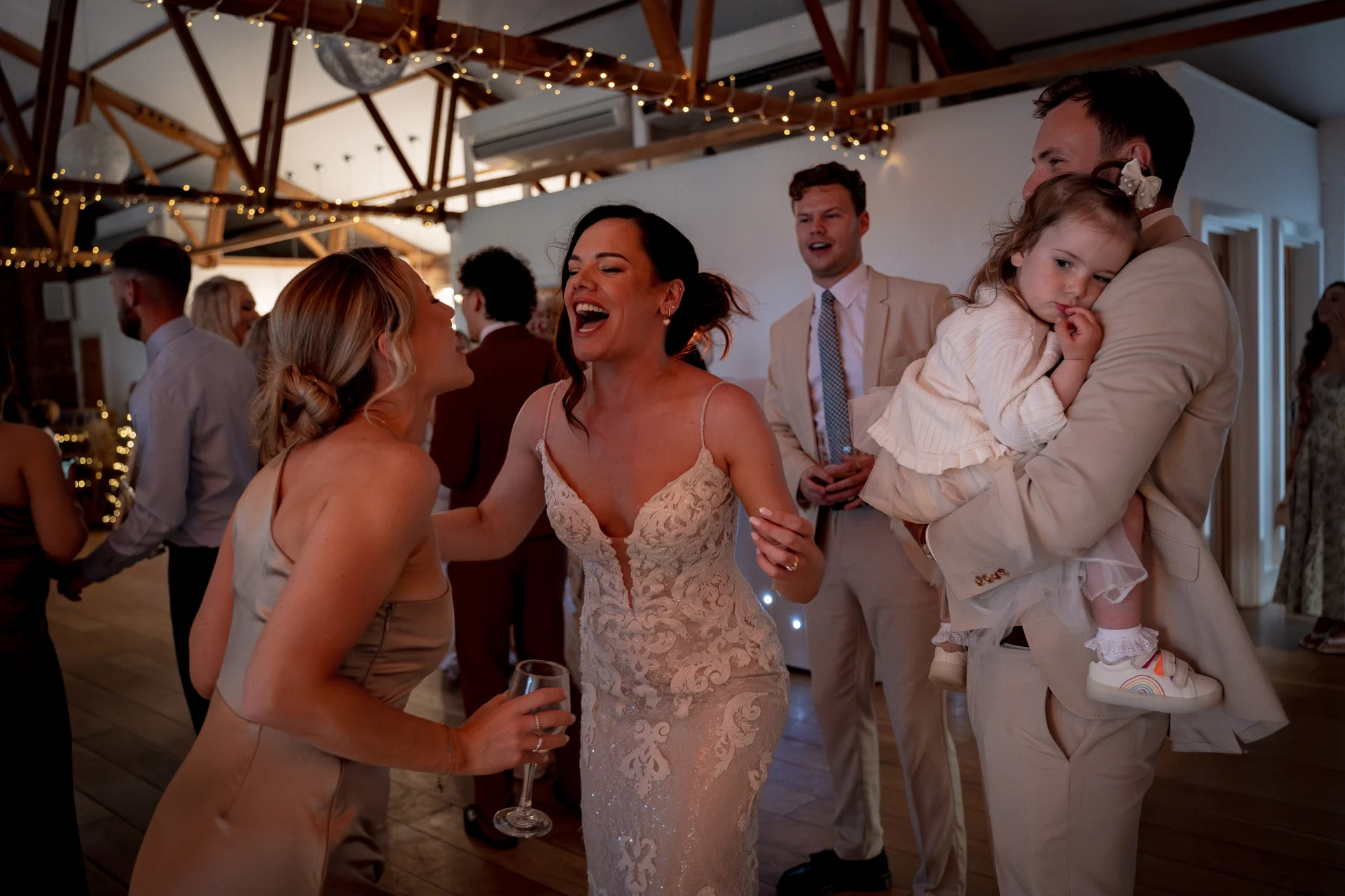 A bride in a lace gown laughs joyfully while dancing at her evening reception inside a rustic barn venue illuminated by fairy lights.