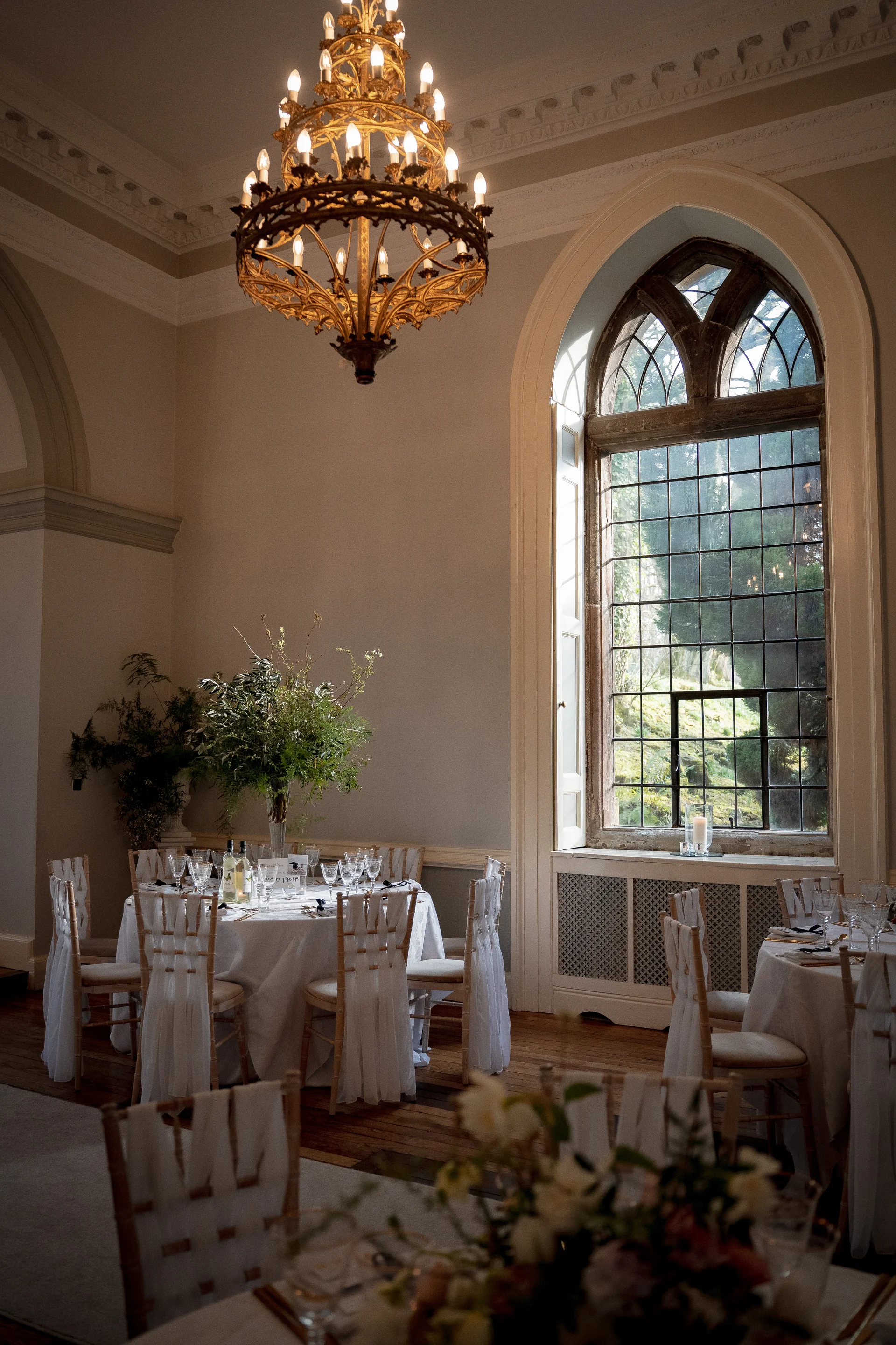 An elegant wedding reception room featuring round tables with white linens, chiavari chairs, and towering arched windows flooded with natural light.