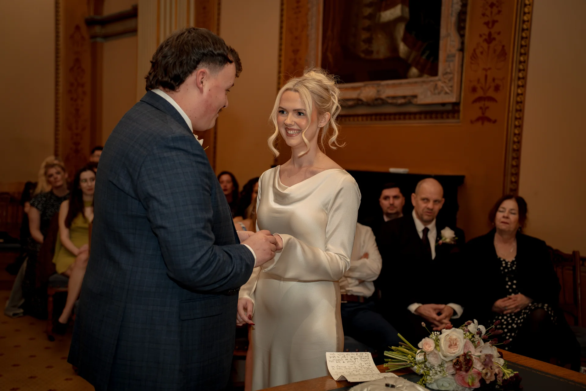 The bride and groom exchange rings during their civil ceremony inside an elegant wood-panelled room, surrounded by seated wedding guests.