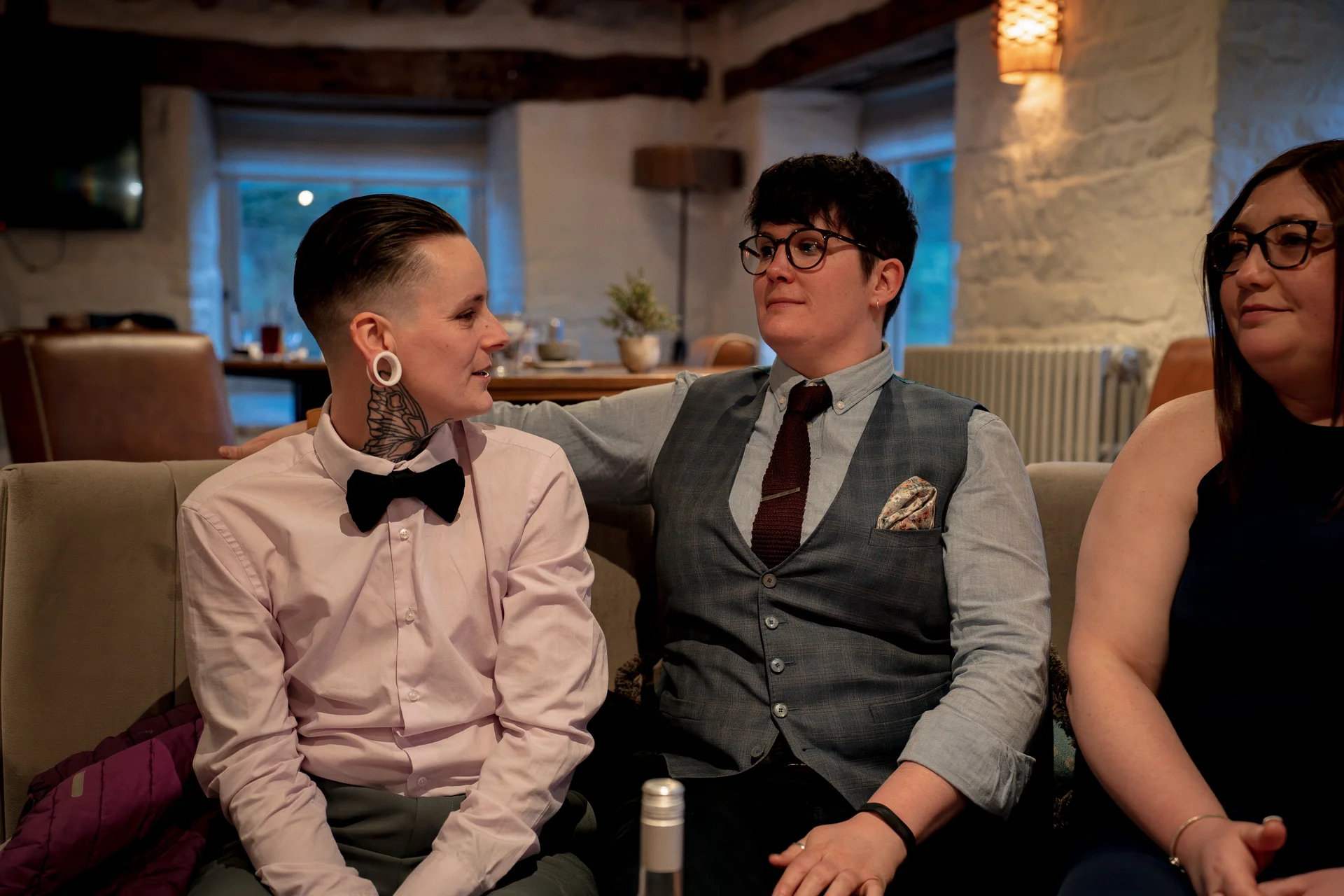 A same-sex couple and a guest sit comfortably together on a sofa inside a rustic venue with exposed brick walls and wooden beams.