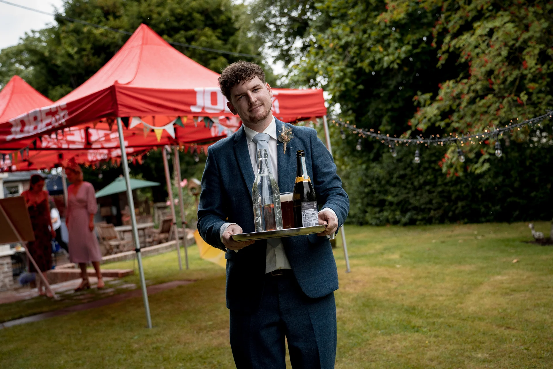 A smiling man in a blue suit holds a tray with bottles and a glass during an outdoor festival wedding with red marquees in the background.