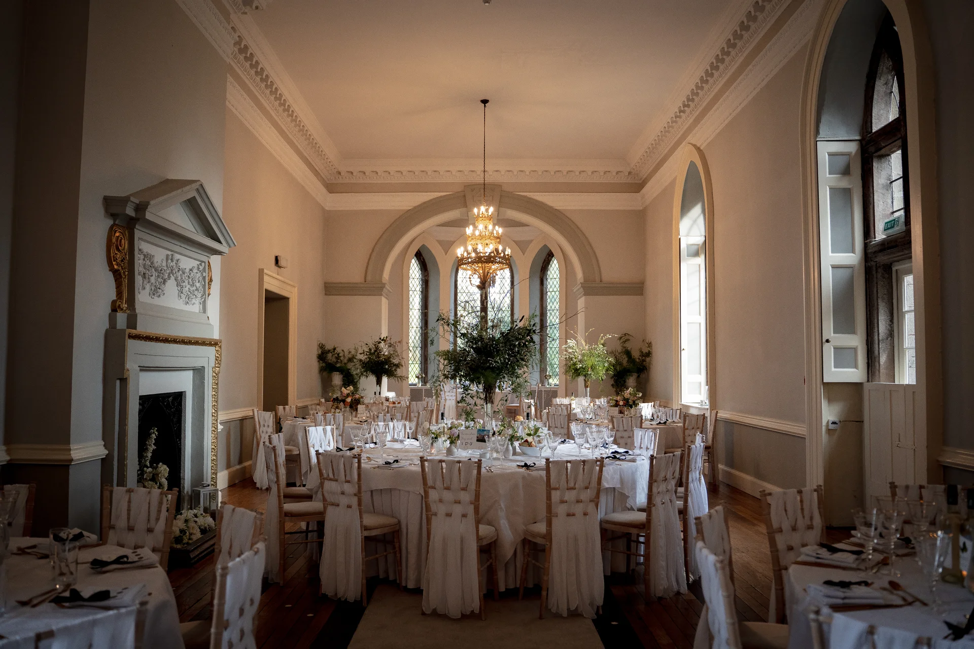 An elegant wedding breakfast reception set up inside a grand manor house ballroom featuring high ceilings, tall arched windows, and round tables with white linens.