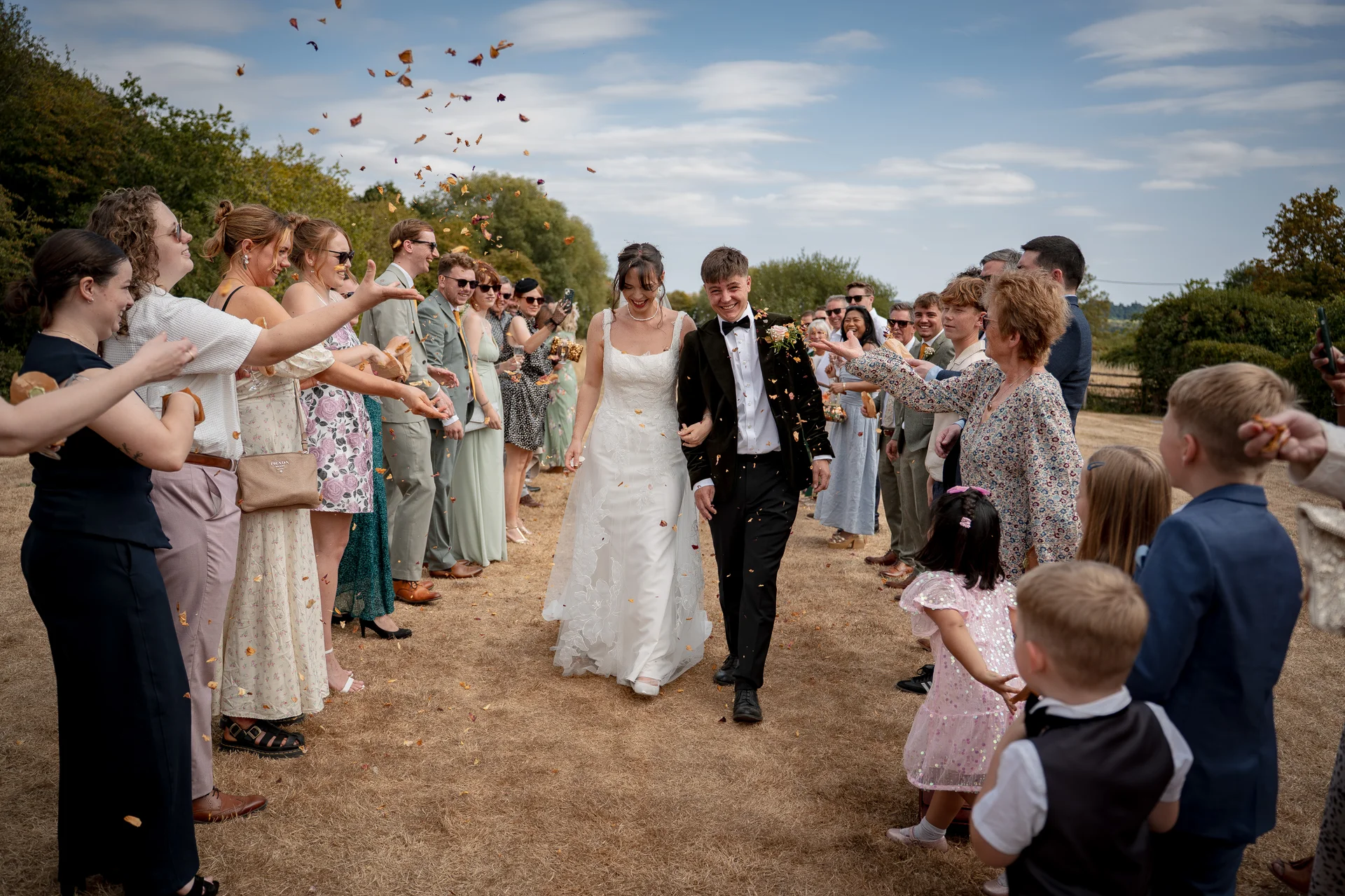 Newlyweds walk happily through a cheering crowd of guests who are showering them with colourful petal confetti during an outdoor daytime wedding.