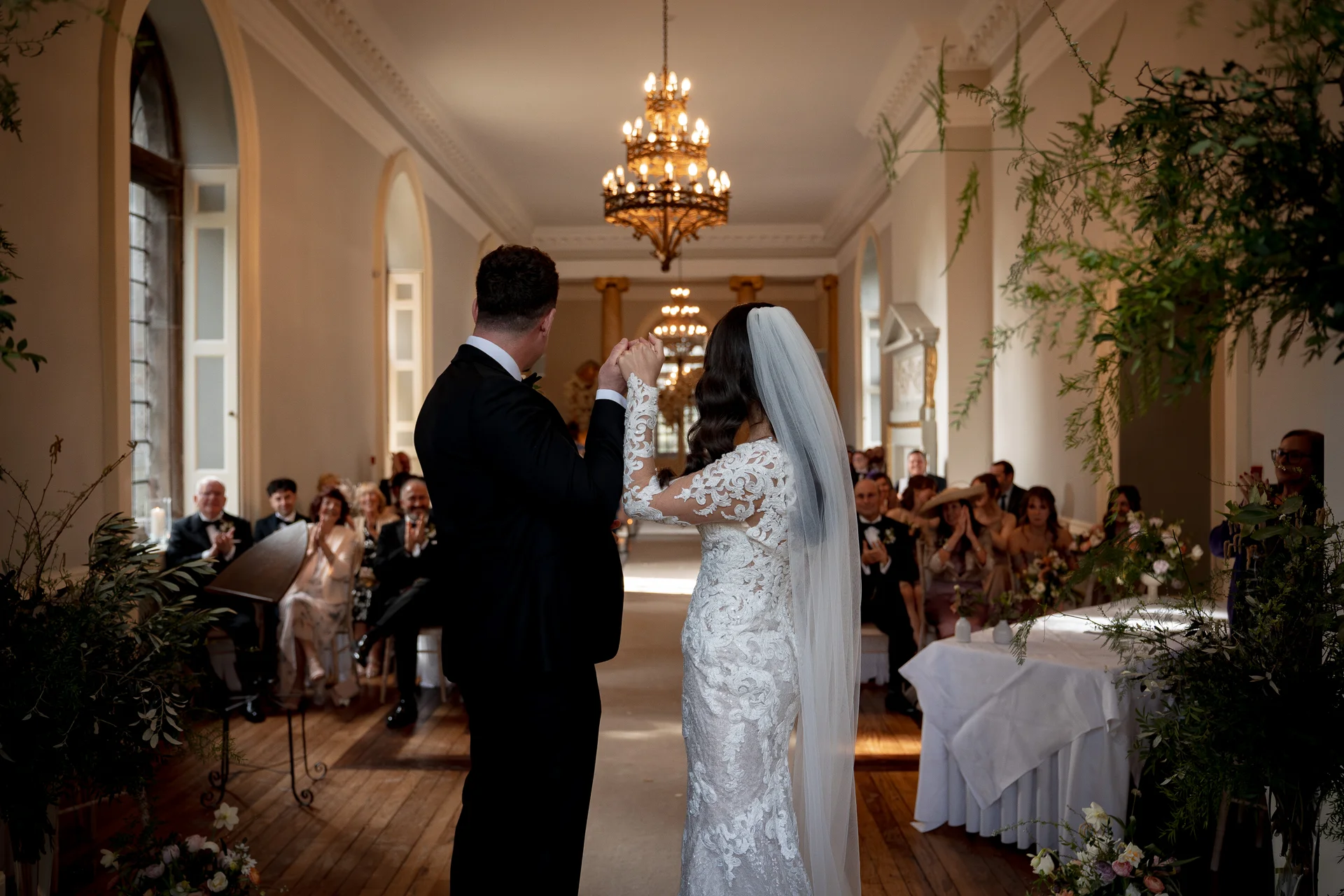 A newly married couple stands hand in hand facing away from the camera in a grand, sunlit hall as seated guests applaud.