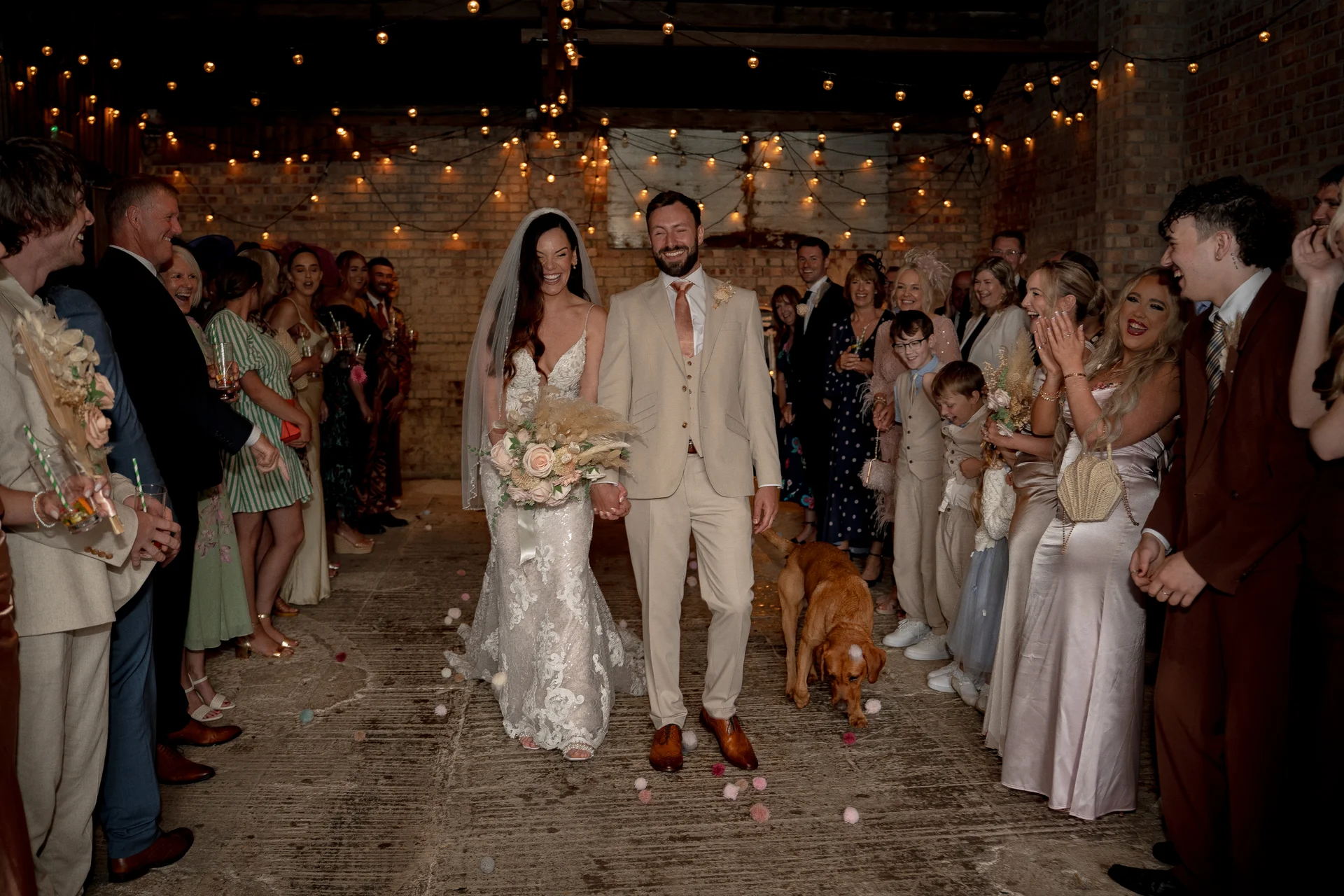 A newlywed couple laughing as they walk through a cheering crowd of wedding guests inside a rustic brick barn adorned with string lights, accompanied by a dog on a leash.