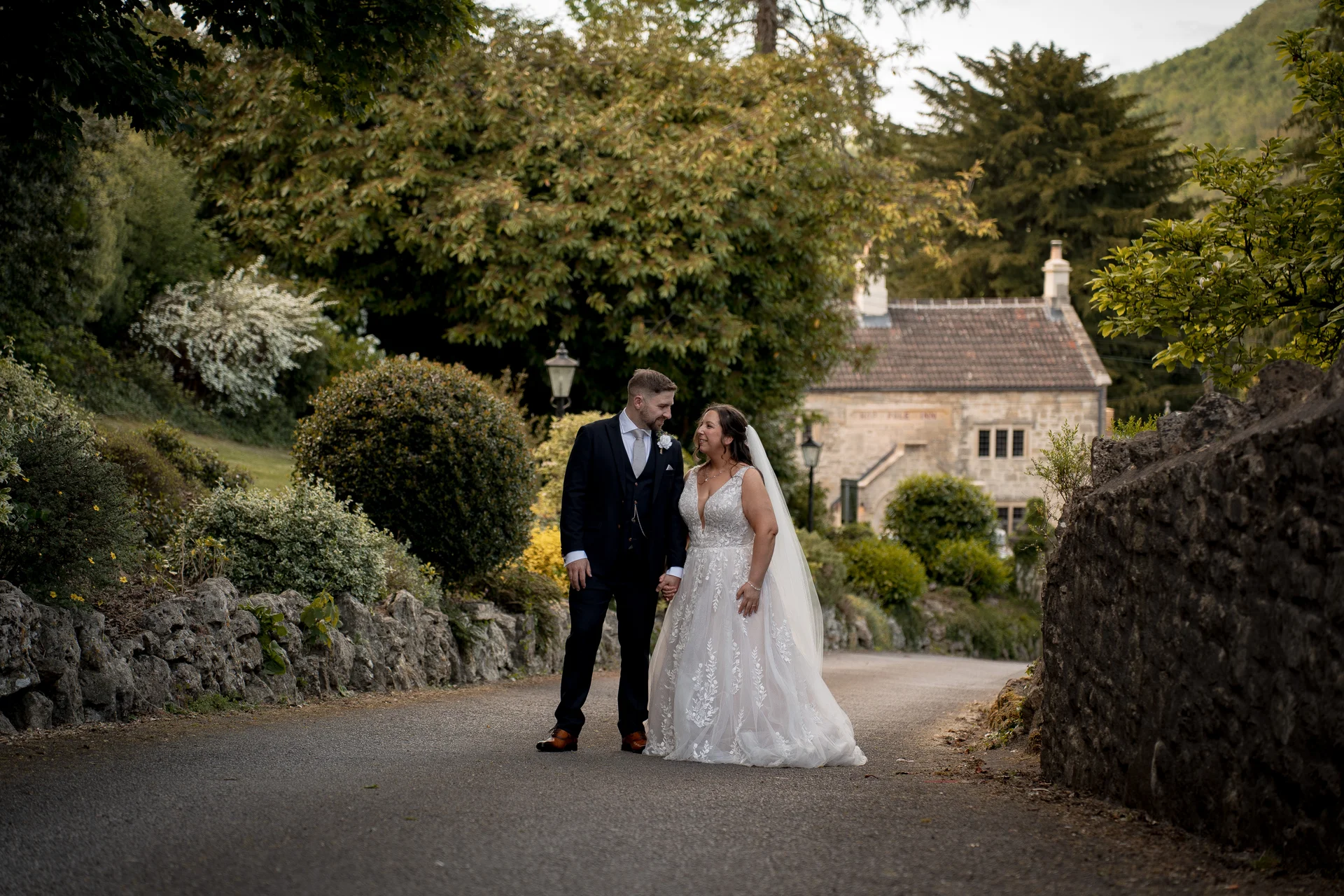 A newlywed couple holds hands and walks along a scenic country path framed by stone walls, mature trees, and a historic stone building in the background.