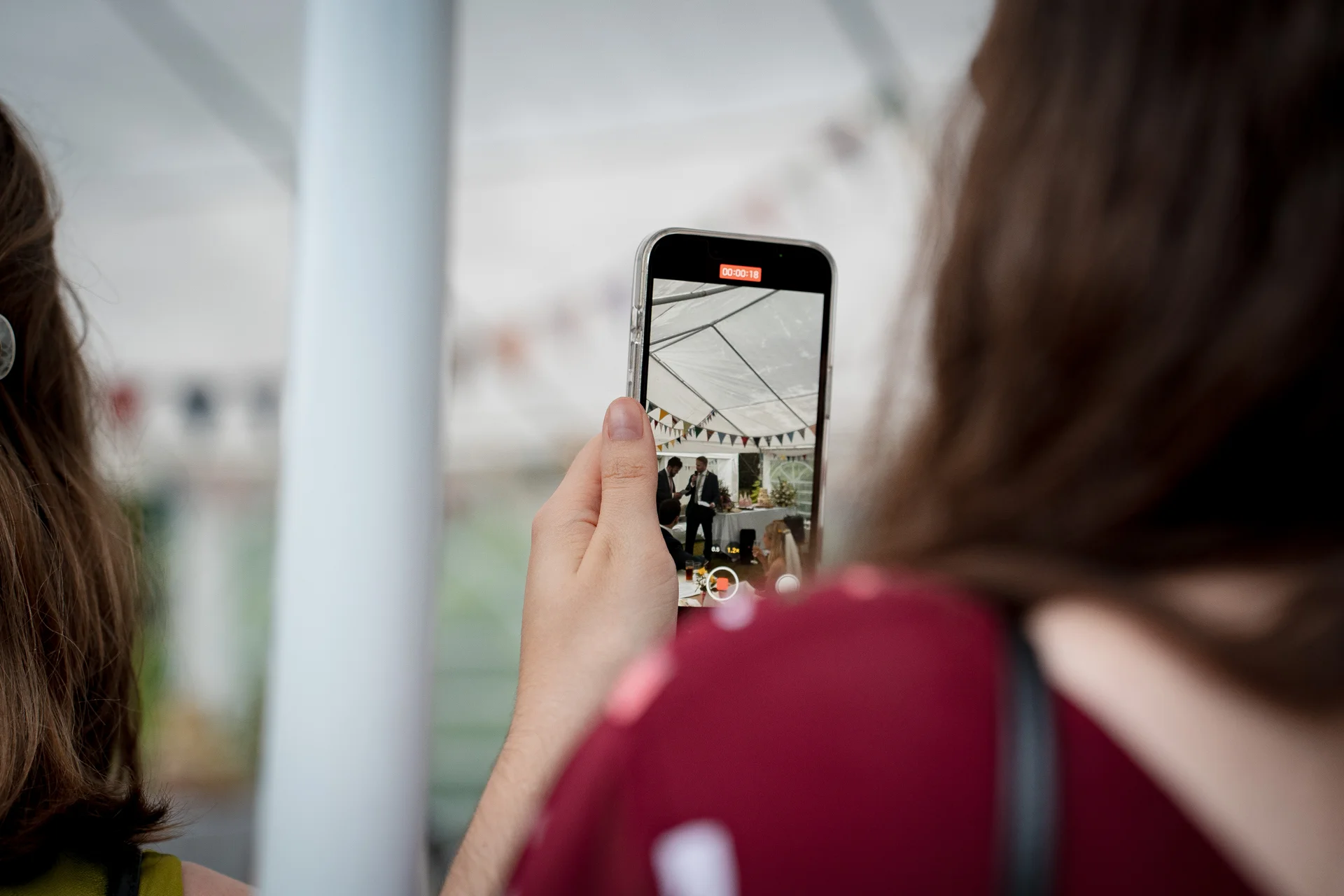 An over the shoulder shot of a wedding guest using a smartphone to record a video inside a marquee decorated with bunting.