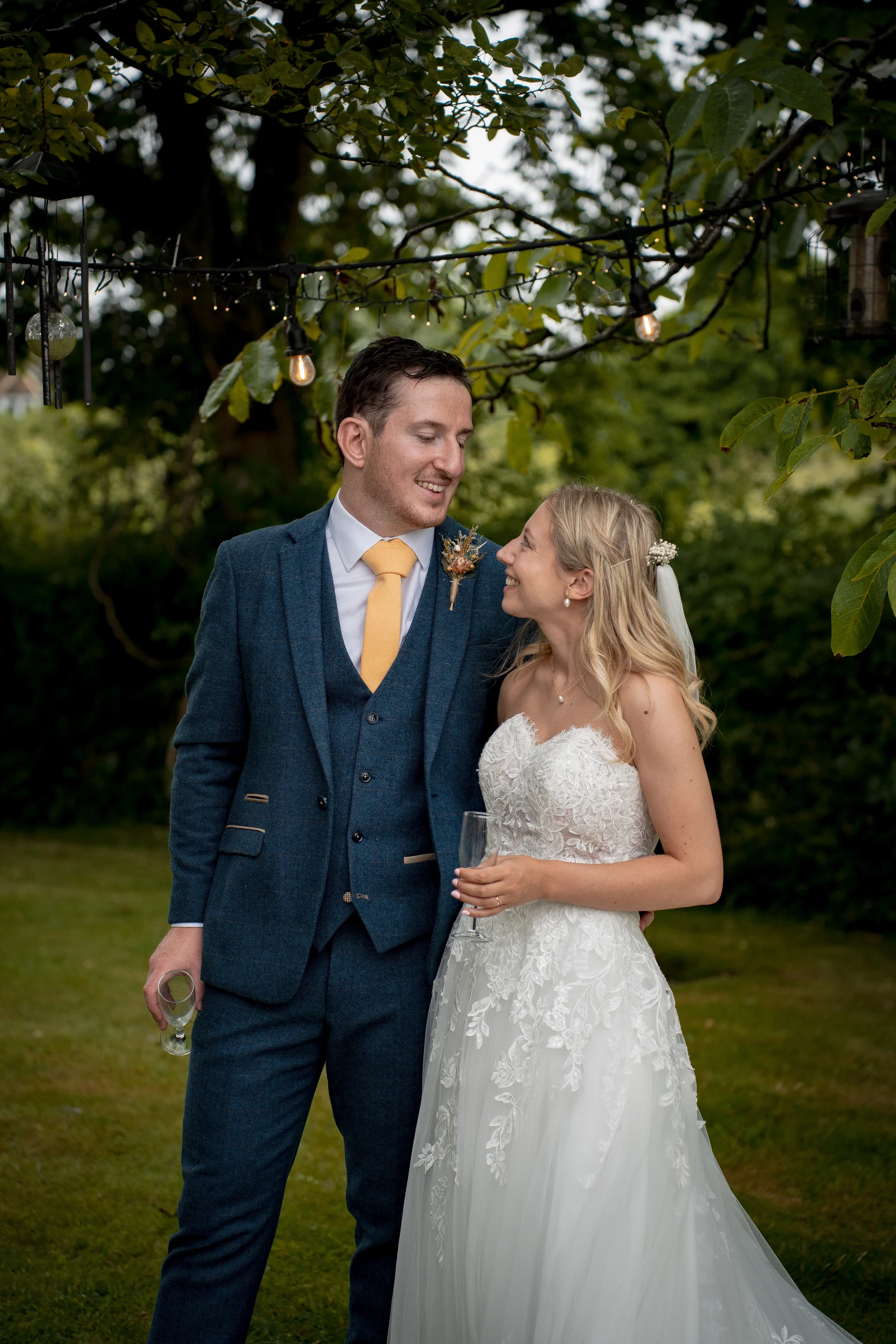 A smiling bride and groom share a loving glance while standing outdoors in a lush garden beneath festoon lighting, holding champagne glasses.