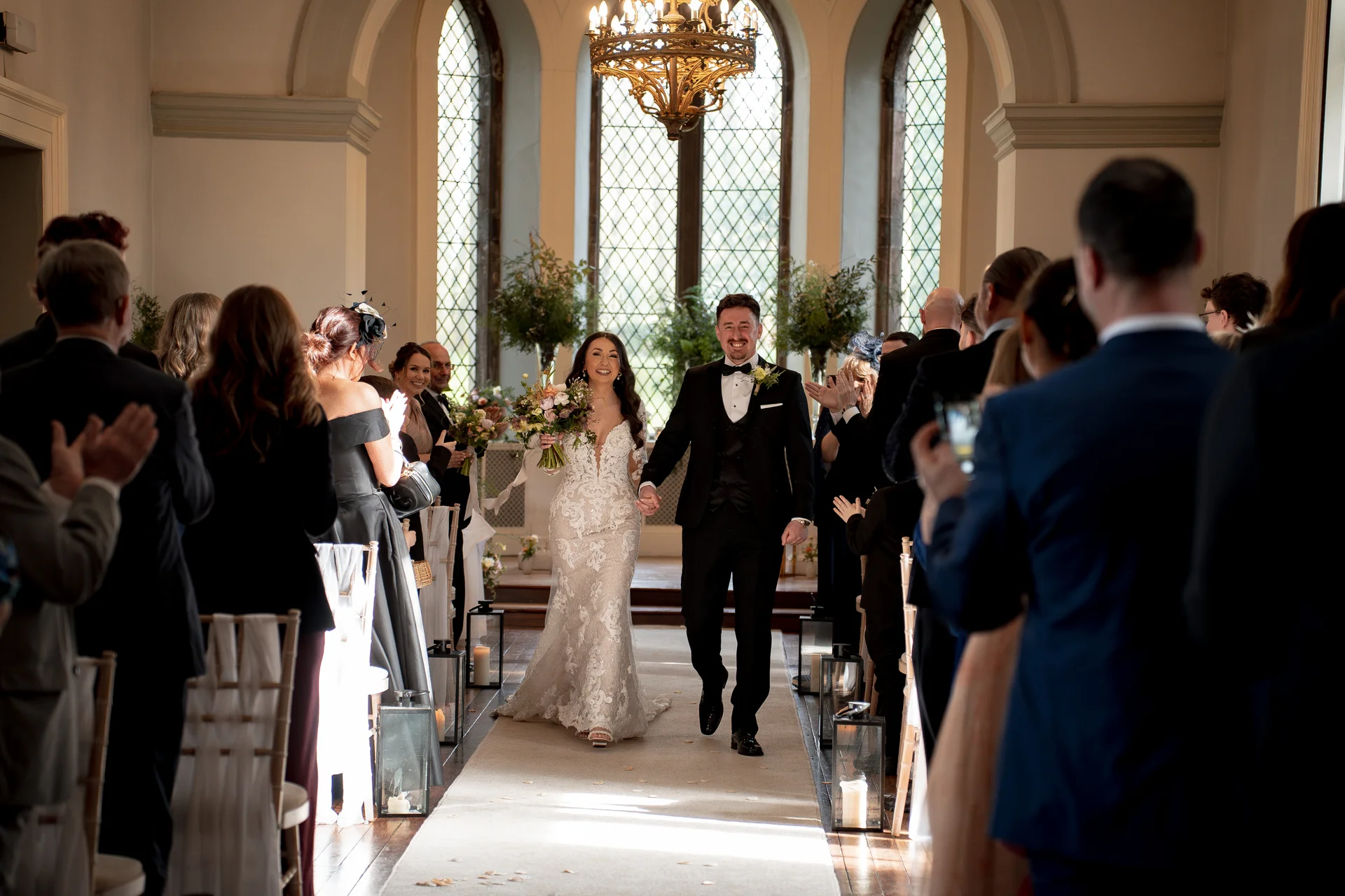 The newlyweds walk hand in hand down the aisle smiling joyfully as guests cheer and applaud in a historic venue with tall arched windows.
