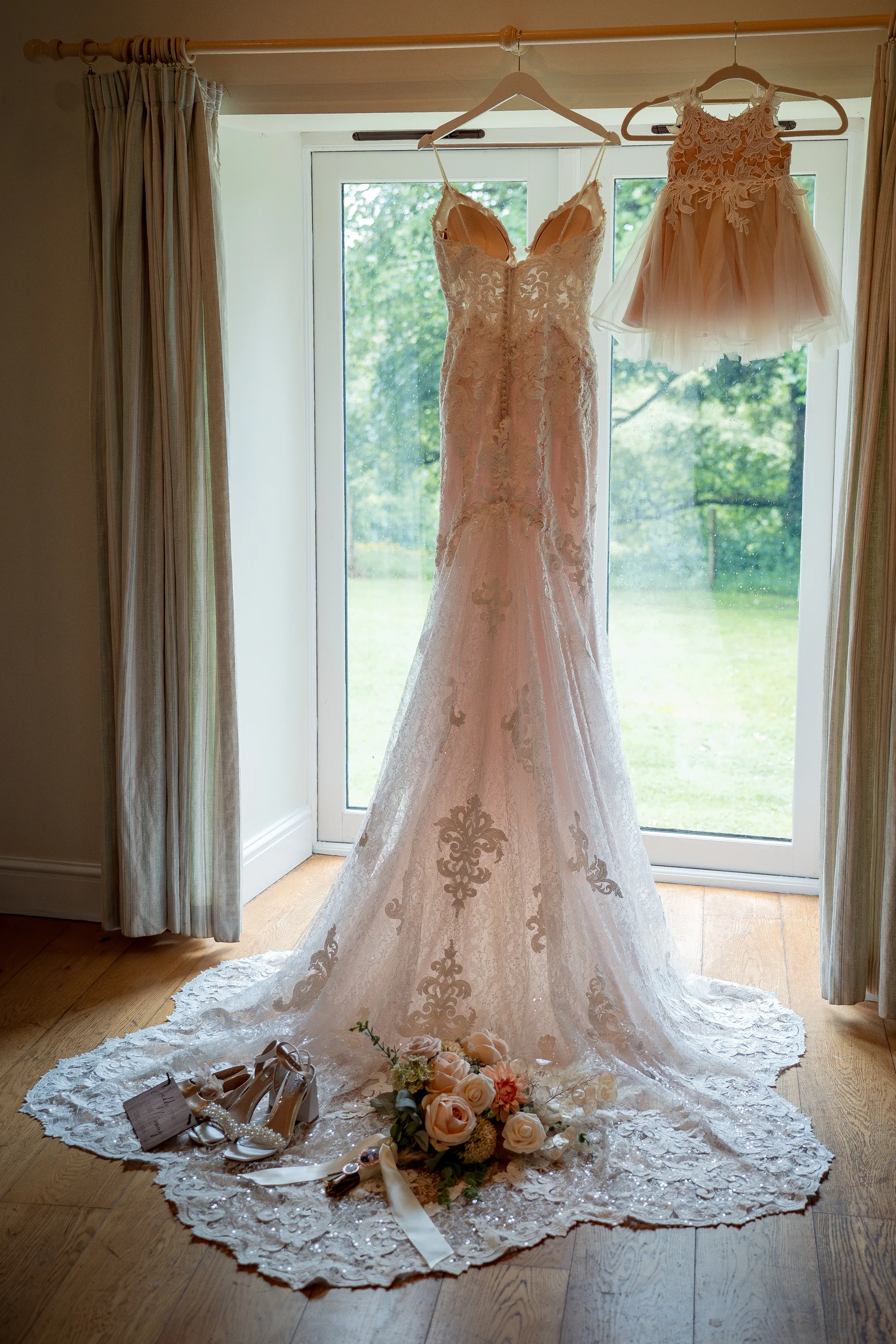 A stunning lace wedding gown and a matching flower girl dress hang gracefully in front of a bright window, with bridal shoes and a delicate floral bouquet arranged on the wooden floor below.