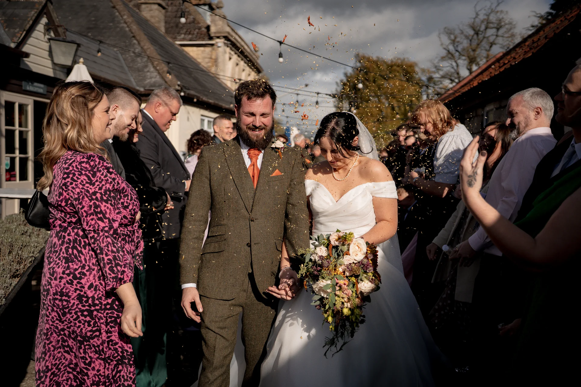 A smiling bride and groom walk hand in hand down an outdoor aisle while wedding guests shower them with golden confetti.