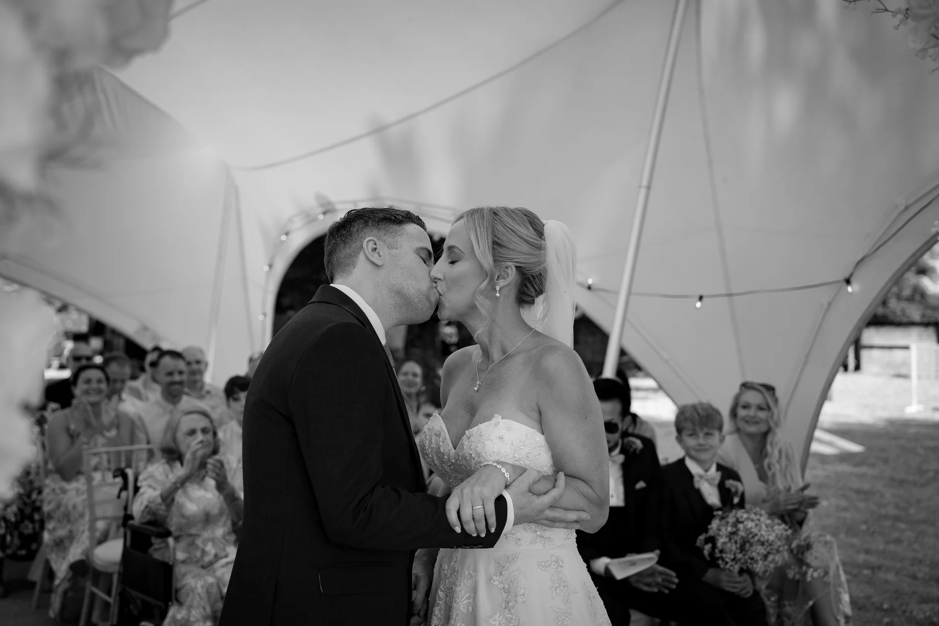 A newlywed couple share their first kiss beneath a white stretch tent canopy during an outdoor wedding ceremony, with seated guests applauding in the background.