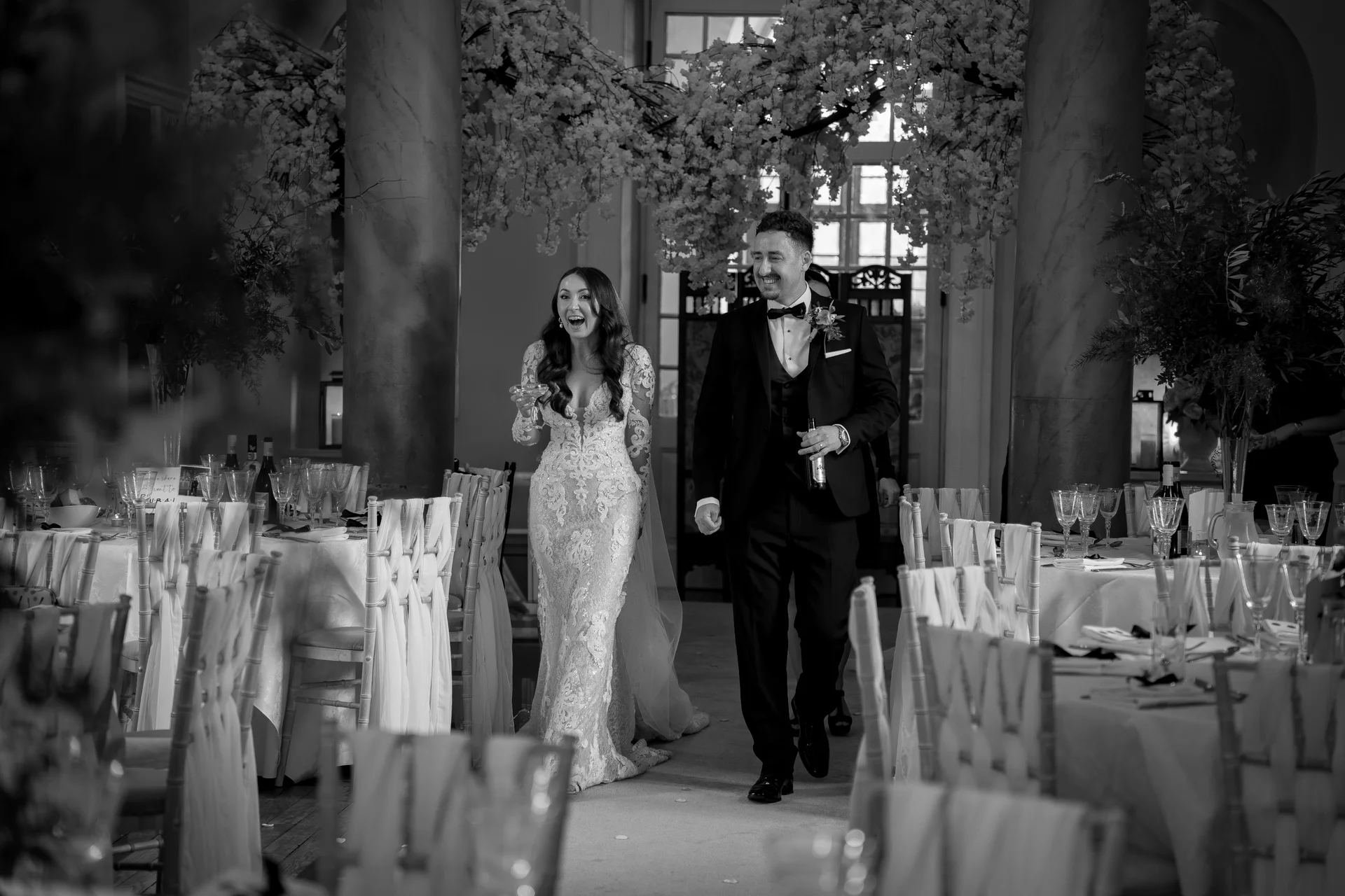 A joy-filled newlywed couple makes their grand entrance into a sunlit, elegantly decorated reception room flanked by dressed dining tables.