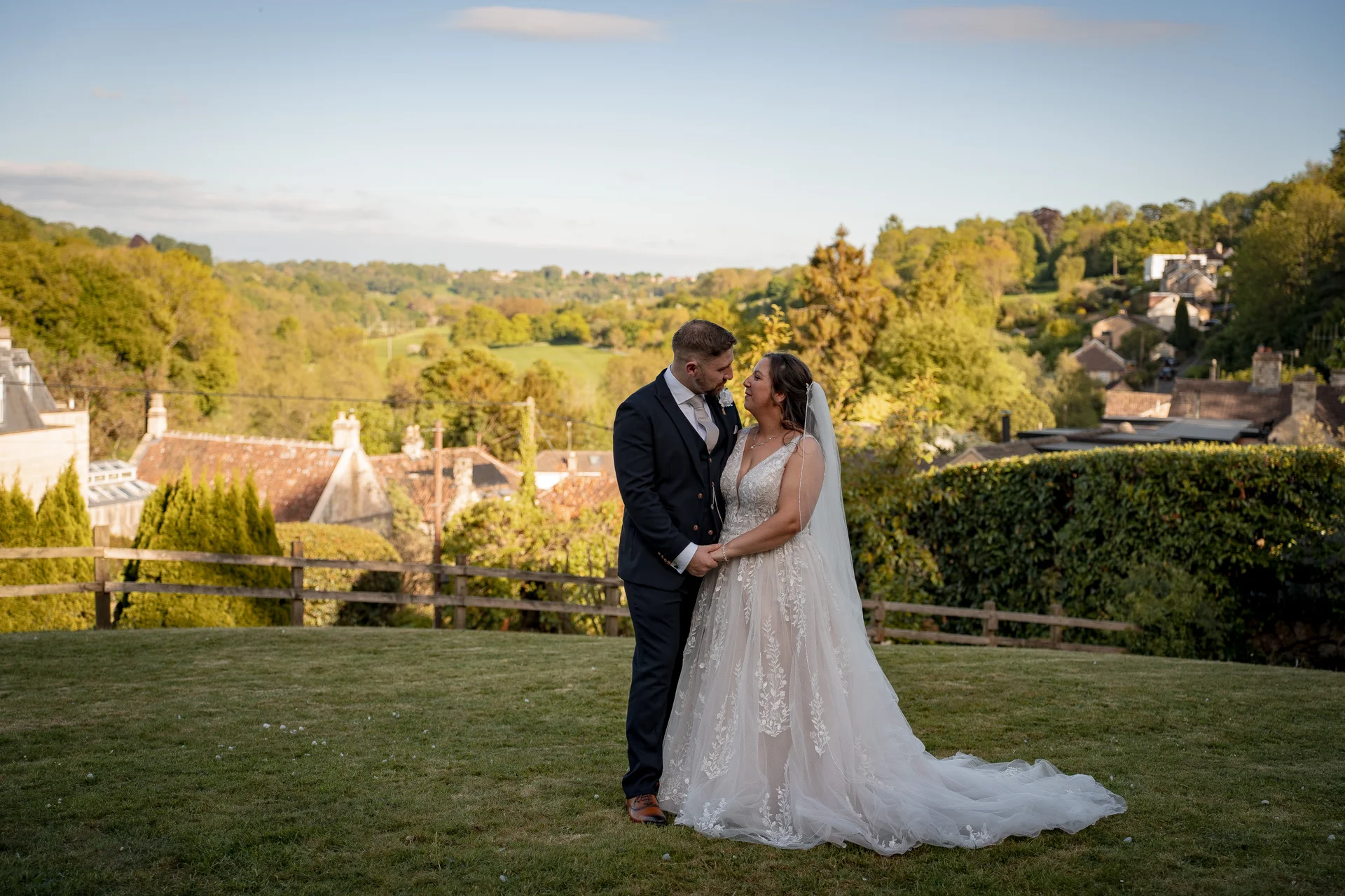 A newlywed couple stands hand-in-hand on a grassy lawn during golden hour, with a scenic backdrop of rolling hills and village rooftops.