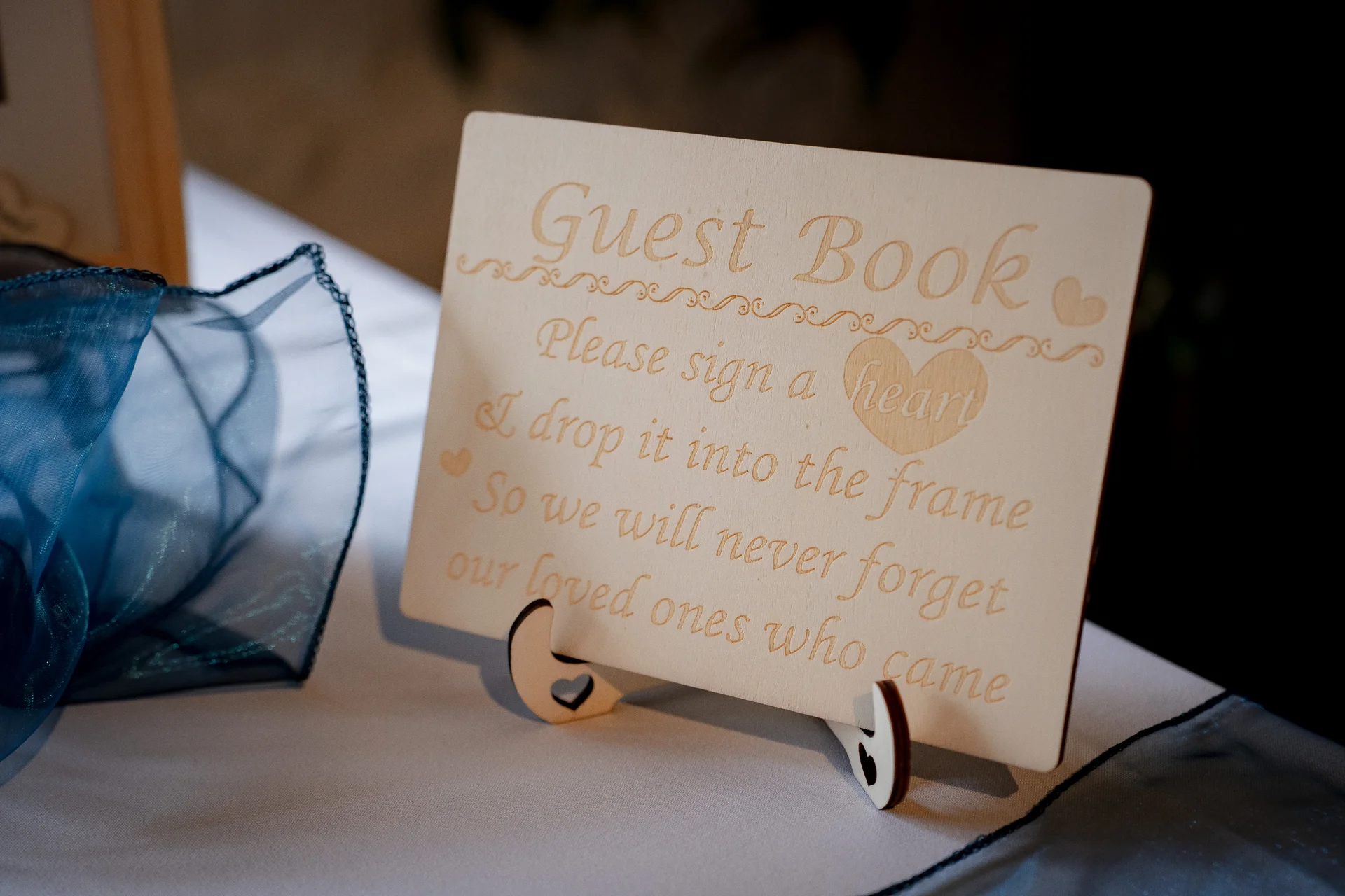 An engraved wooden sign on a tabletop requests guests to sign a heart and drop it into the frame for the wedding guest book.