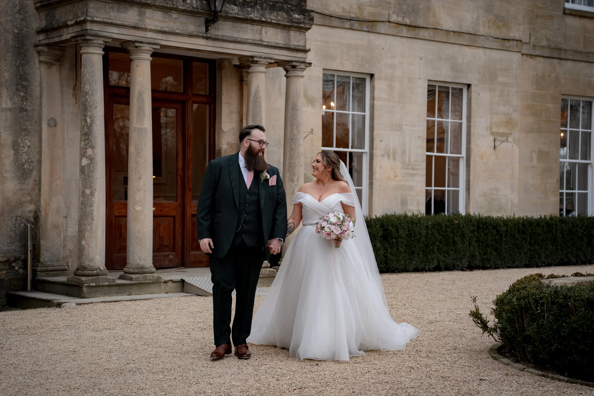 A newlywed couple holds hands and smiles at each other outside the grand stone entrance of a country house venue.