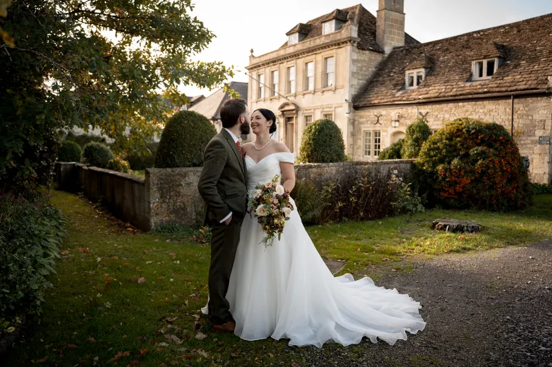 A bride holding an autumn bouquet and a groom in a tweed suit share a tender moment outside a historic country house at golden hour.